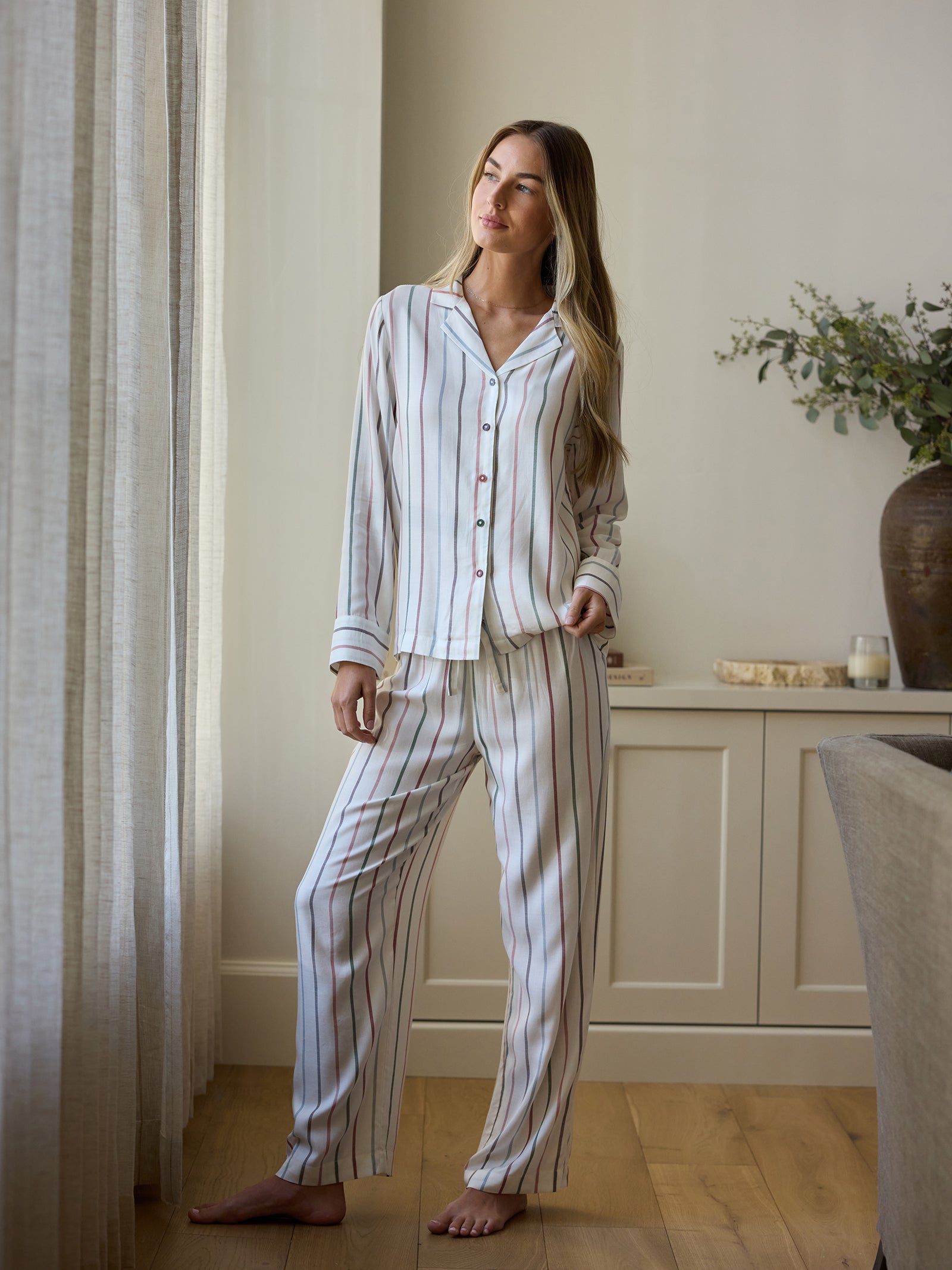 A person stands by a window wearing Cozy Earth's Women's Soft Woven Pajama Pant. The room has a light, airy feel with plants and decor on a shelf in the background. 