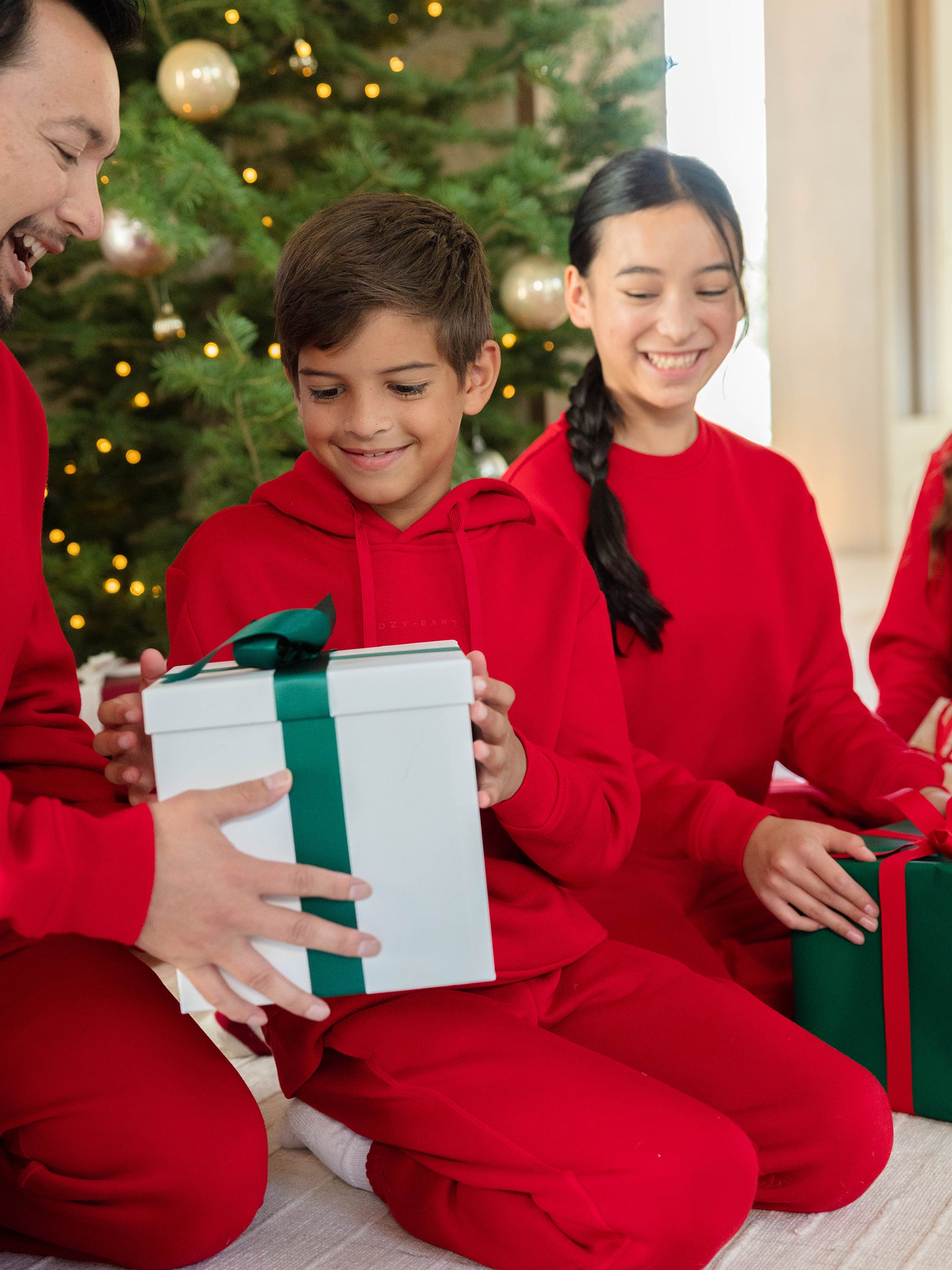 A family in matching red outfits sits by a Christmas tree, including a boy wearing the Kid's CityScape Hoodie from Cozy Earth. He receives a gift wrapped in white paper with a green ribbon from an adult, adding to the warm, festive atmosphere with ornaments and lights beautifully illuminating the background. 