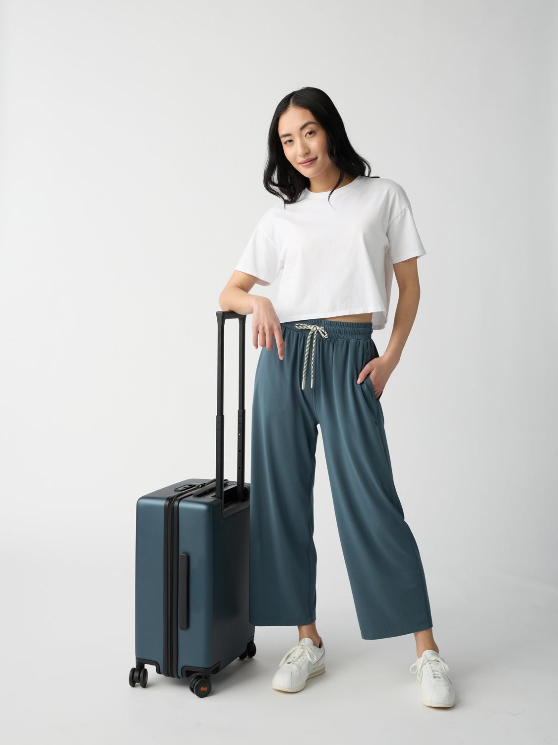 A young woman with long dark hair stands smiling in a studio, wearing a white t-shirt, Cozy Earth's Women's Studio Cropped Wide Leg Pant in blue, and white sneakers. She holds a blue suitcase against a plain white background. 