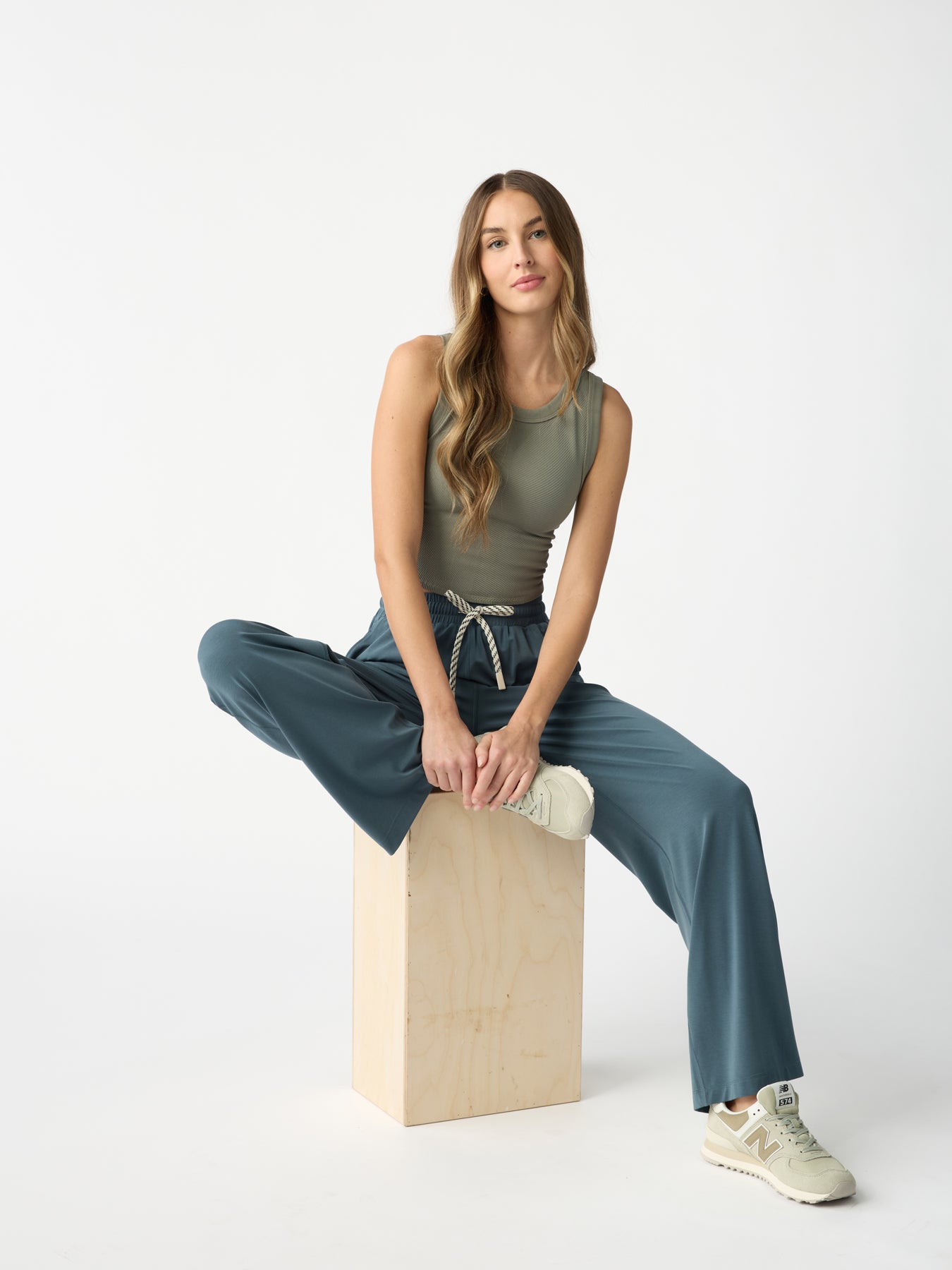 A woman with long hair sits on a wooden box against a white backdrop, wearing a sleeveless green top, Cozy Earth's Women's Studio Wide Leg Pant in blue, and beige sneakers. One leg is bent while the other hangs down for a relaxed pose. 