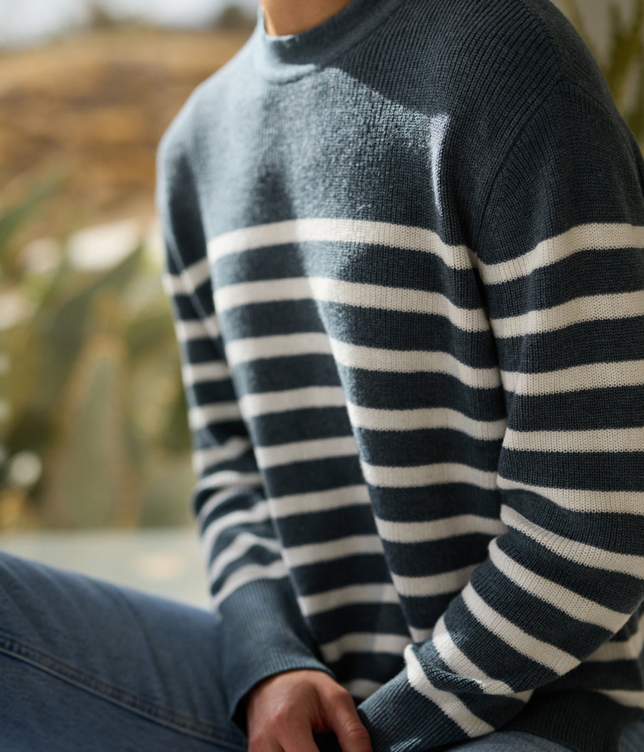 A person in a Women's Rowan Sweater from Cozy Earth, featuring blue and white stripes, paired with blue jeans, is seated in a brightly-lit room. The focus remains on their upper body and attire against a softly blurred backdrop. 