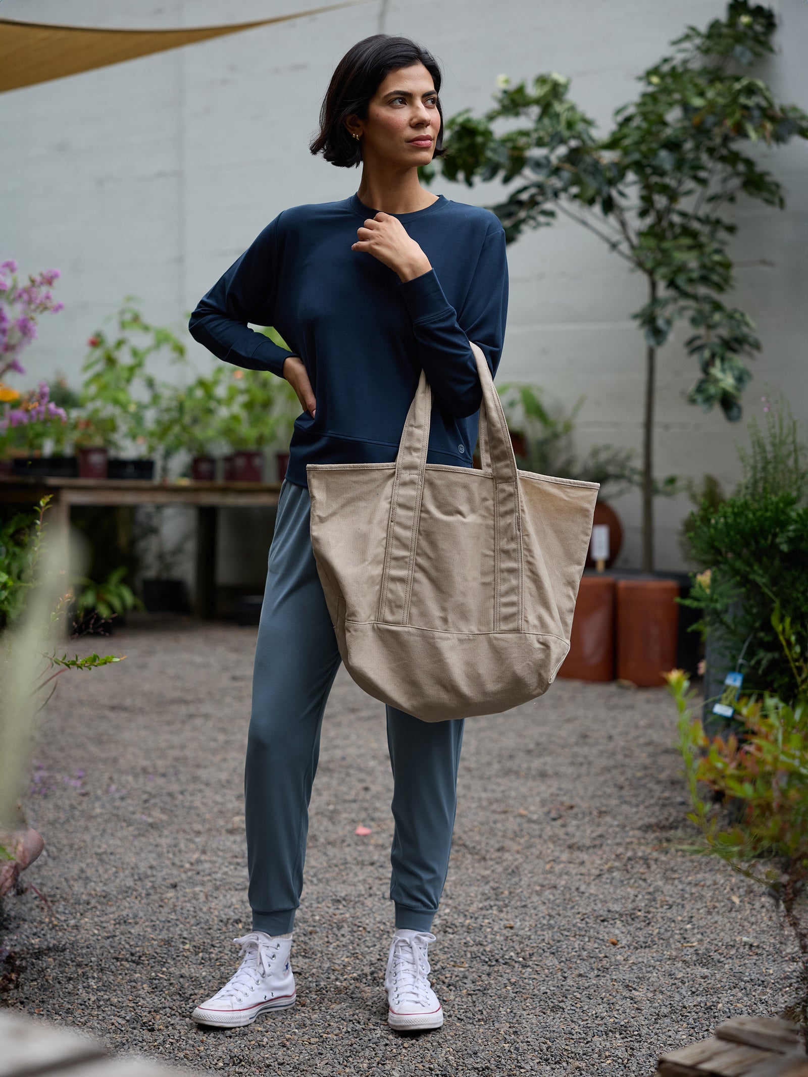 A person stands outdoors on a gravel path, holding the strap of a large beige tote bag. They are wearing a navy blue long-sleeve shirt, Cozy Earth's Women's Studio Jogger in light blue, and white sneakers. The surroundings include potted plants and a tree in a garden setting. 