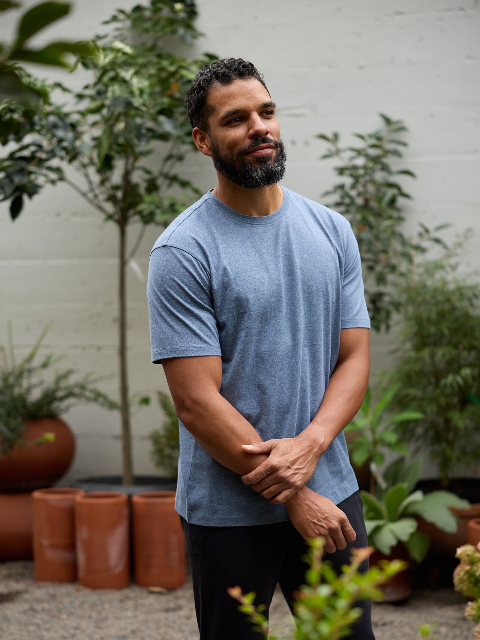 A man with a short beard and short curly hair stands outdoors in front of a garden, wearing a light blue Cozy Earth Men's All Day Tee and dark pants. He is looking slightly to the side with his hands loosely clasped in front of him. Potted plants and trees are in the background. 