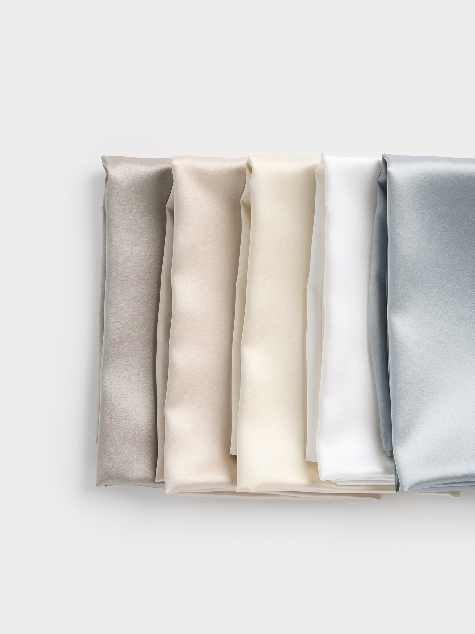 A vertical display of neatly folded Silk Pillowcases from Cozy Earth is shown against a light backdrop, featuring five colors: gray, beige, cream, white, and light blue. The fabric looks smooth and silky. 