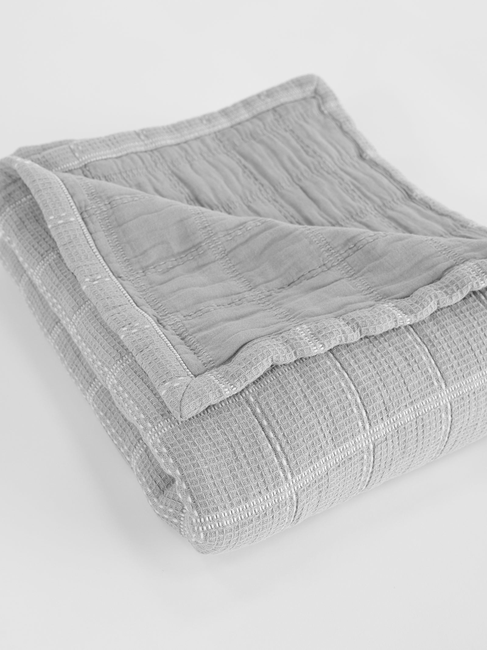 The Waffle Windowpane Blanket by Cozy Earth, neatly folded in a light gray tone, lies on a flat surface. The blanket showcases its textured weave and detailed stitching, with one corner elegantly flipped over to reveal the intricate underside. 