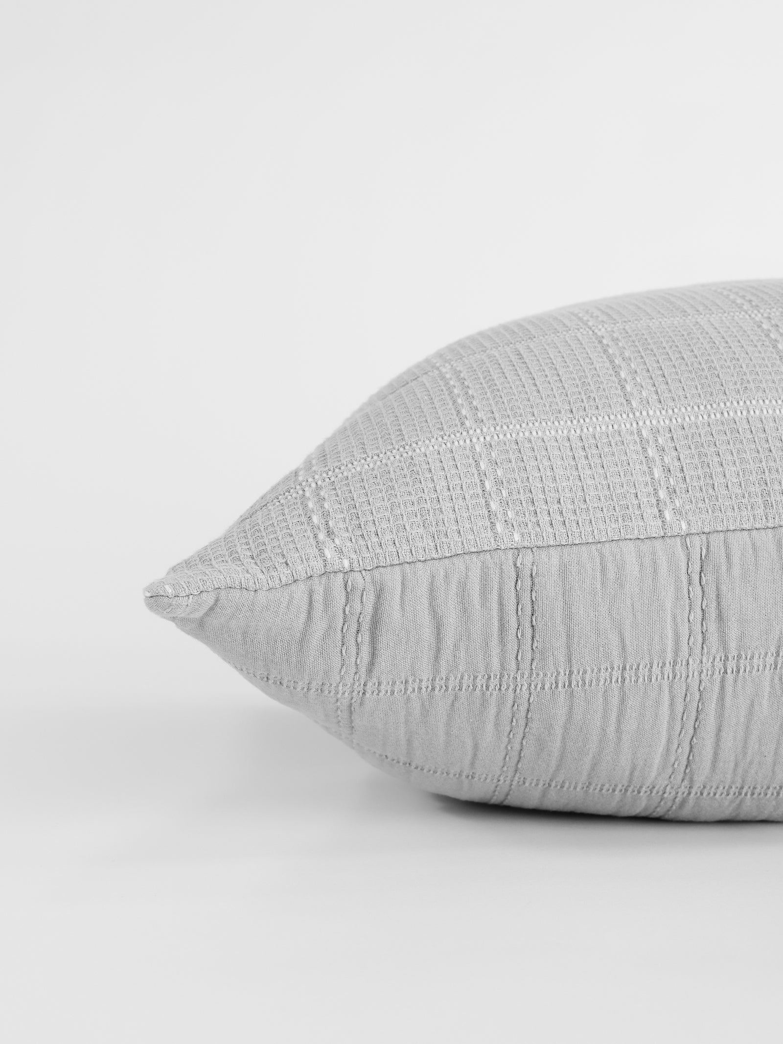 A close-up of the Waffle Windowpane Pillow by Cozy Earth reveals its gray, textured, grid-patterned fabric. The pillow is captured from a side angle, showcasing its soft and slightly plump appearance against a plain white background. 