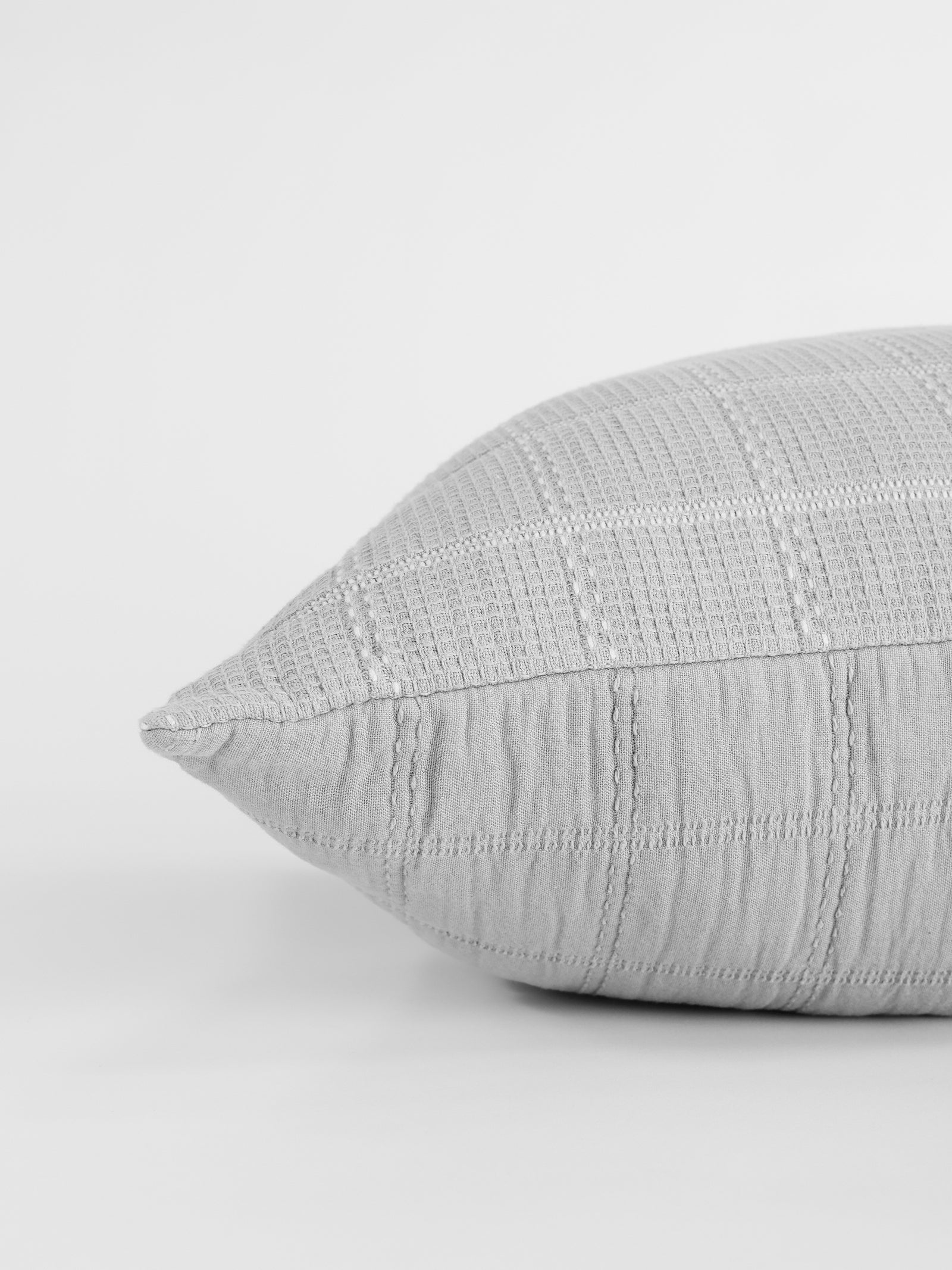 A close-up view of the Cozy Earth Waffle Windowpane Pillow, featuring a gray textured, stitched grid pattern, set against a plain, light-colored background. 