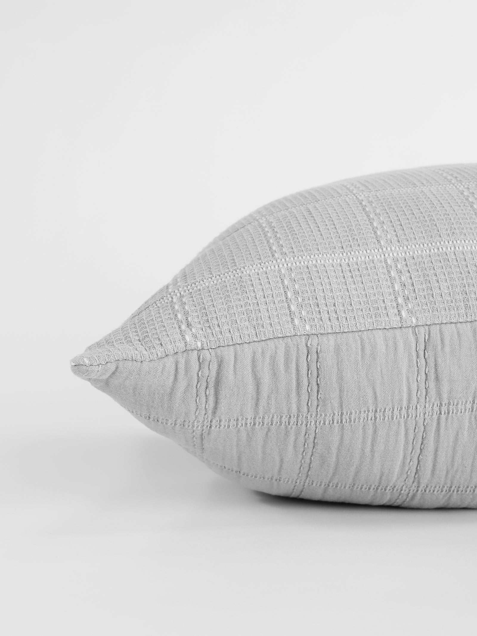 Close-up of the corner of a light gray, textured Waffle Windowpane Pillow by Cozy Earth. The pillow features a subtle crisscross pattern with slightly raised lines, giving it a quilted appearance. The plain white background highlights the pillow’s texture and color. 