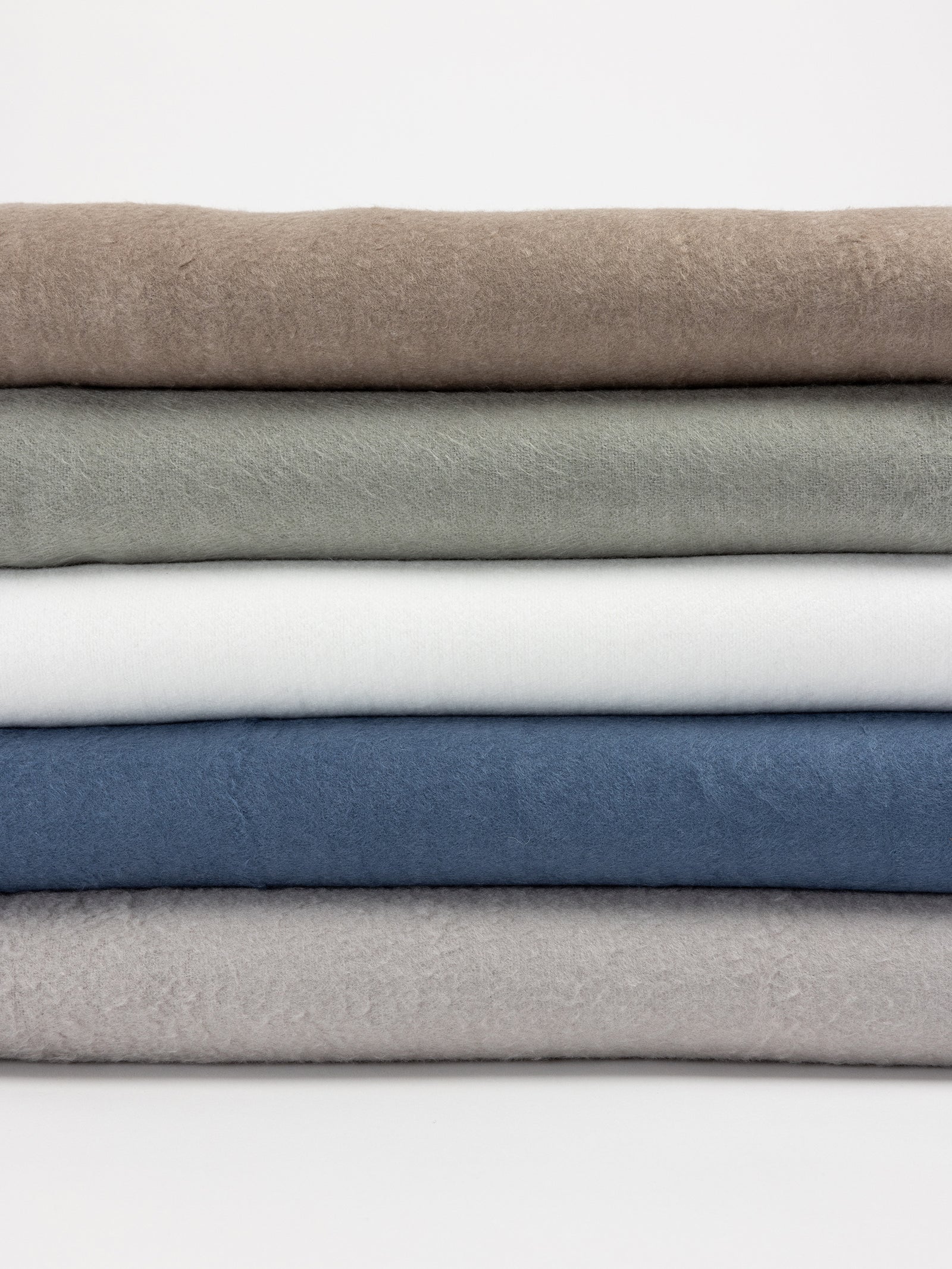 A stack of five Cozy Earth Bamboo Blankets in various colors: beige at the top, followed by green, white, blue, and light gray at the bottom. The blankets have a soft, fuzzy texture and are arranged neatly against a plain white background. 