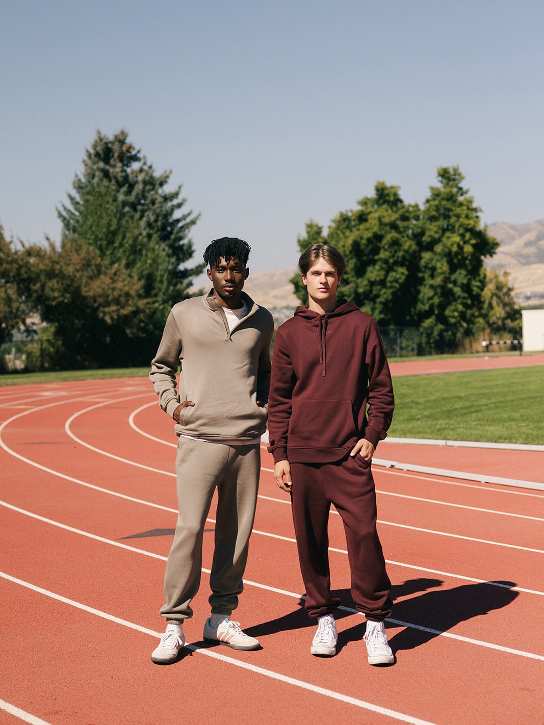 Two people stand on a track field, dressed in casual athletic wear from Cozy Earth. One person is wearing a light grey Men's CityScape Quarter Zip and joggers, while the other sports a maroon Men's CityScape Quarter Zip paired with matching joggers. Both have their hands in their pockets and are looking at the camera, with trees and hills providing a scenic background. 