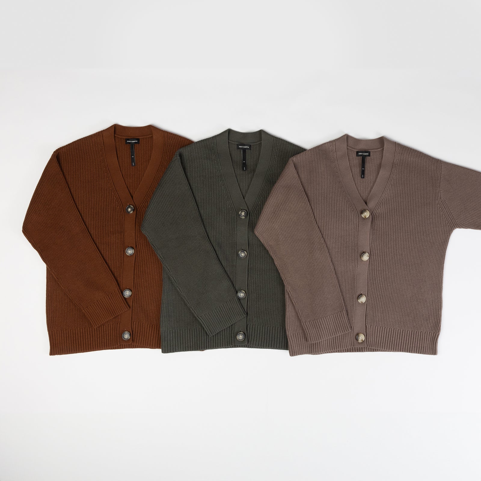 Three Oversized Classic Cardigans from Cozy Earth are laid out flat side by side. The cardigans are in brown, green, and taupe colors, each featuring a V-neck and button closure. 