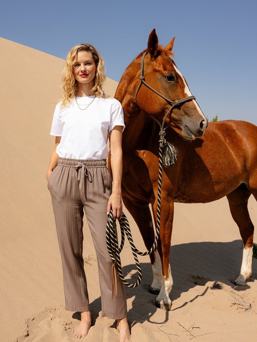A woman with blonde hair, barefoot on sand beside a brown horse, wears Cozy Earth's Women's Sunset Cropped Pant in taupe and a white t-shirt, standing before a sand dune under a clear blue sky. 