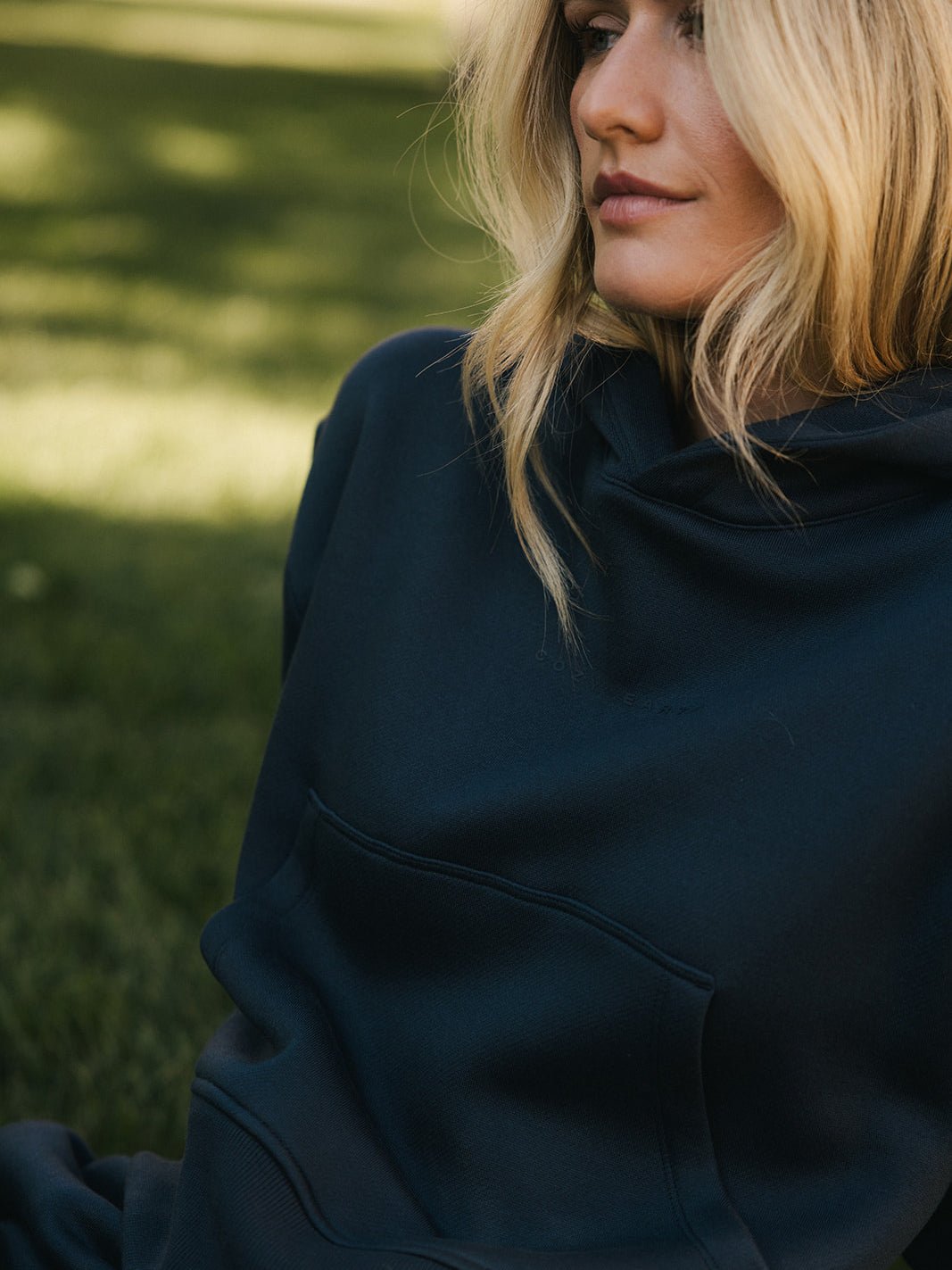 A person with long blonde hair in a Women's CityScape Cropped Hoodie by Cozy Earth is sitting outdoors on the grass. Their face is partially visible, and they appear to be relaxed, enjoying the serene environment. The background is blurred, highlighting a green, grassy area. 