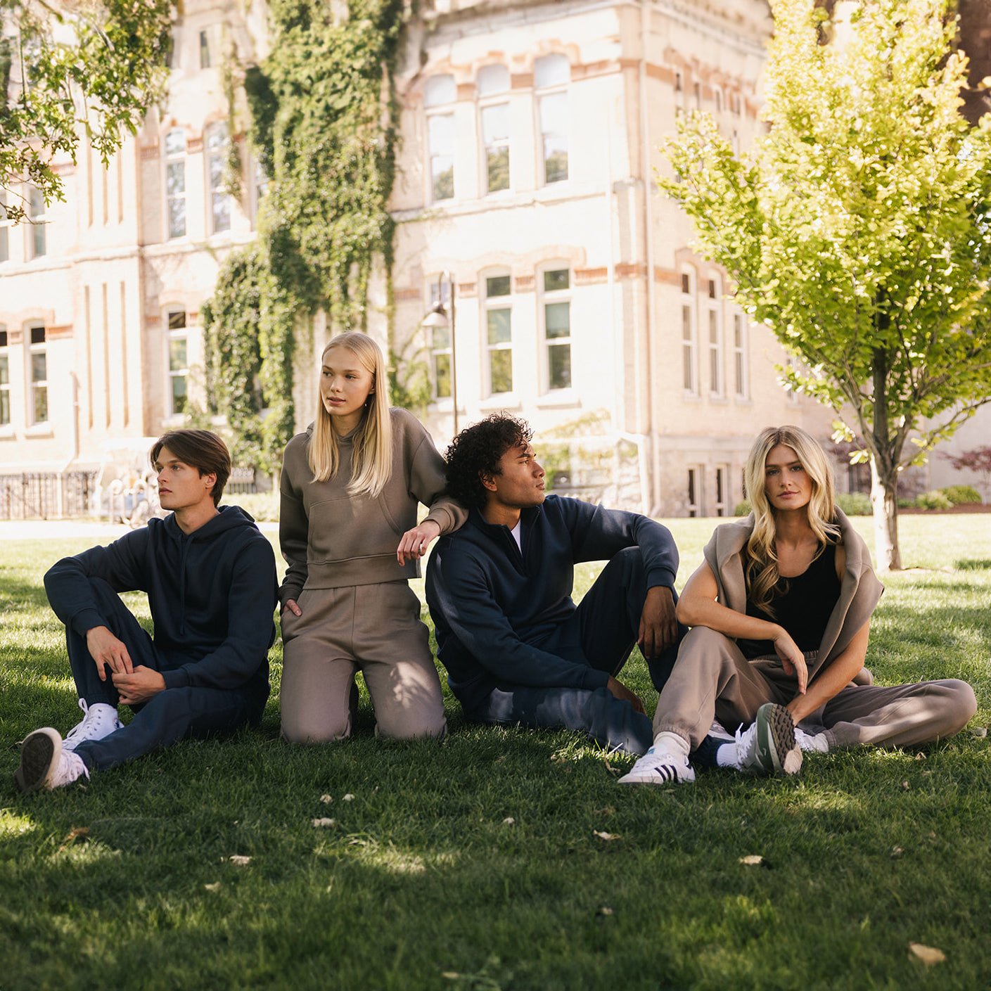 Four individuals sit and lean on each other on a grass lawn in front of a large building with ivy-covered walls. They are dressed in Cozy Earth's Men's CityScape Hoodies in casual, earth-toned colors and look relaxed. Trees with green and yellow leaves add a natural touch to the background. 