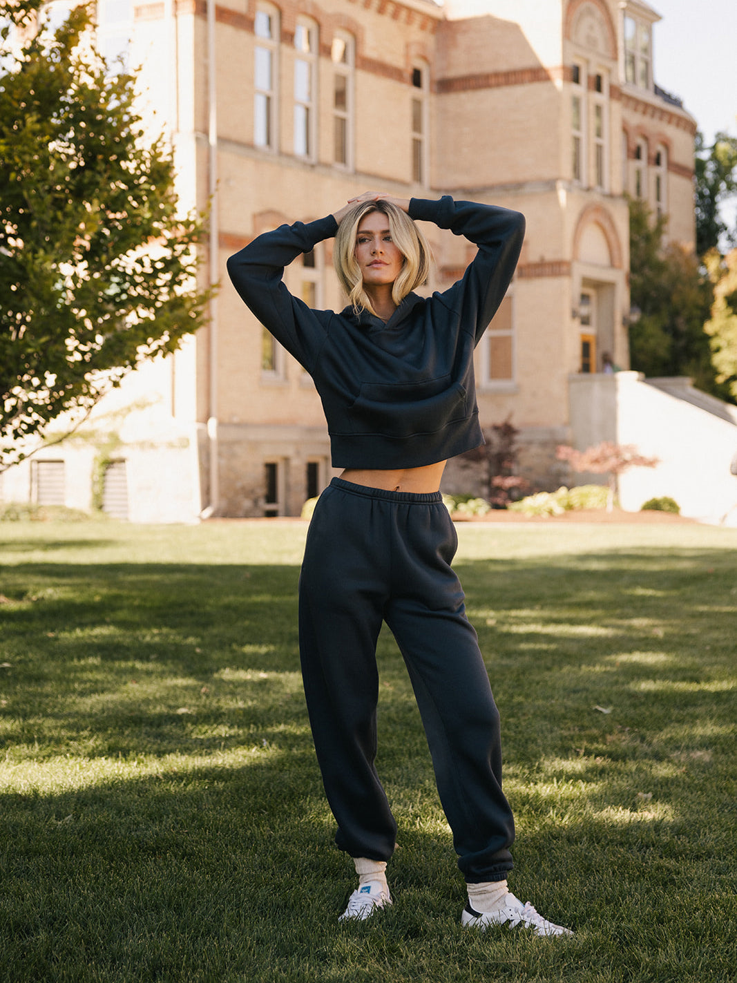 A person wearing a black hoodie and matching Women's CityScape Sweatpants from Cozy Earth stands with hands on their head on a grassy lawn in front of a historic brick building. The person has short blonde hair and is also wearing white sneakers. The scene is in daylight, with trees nearby. 