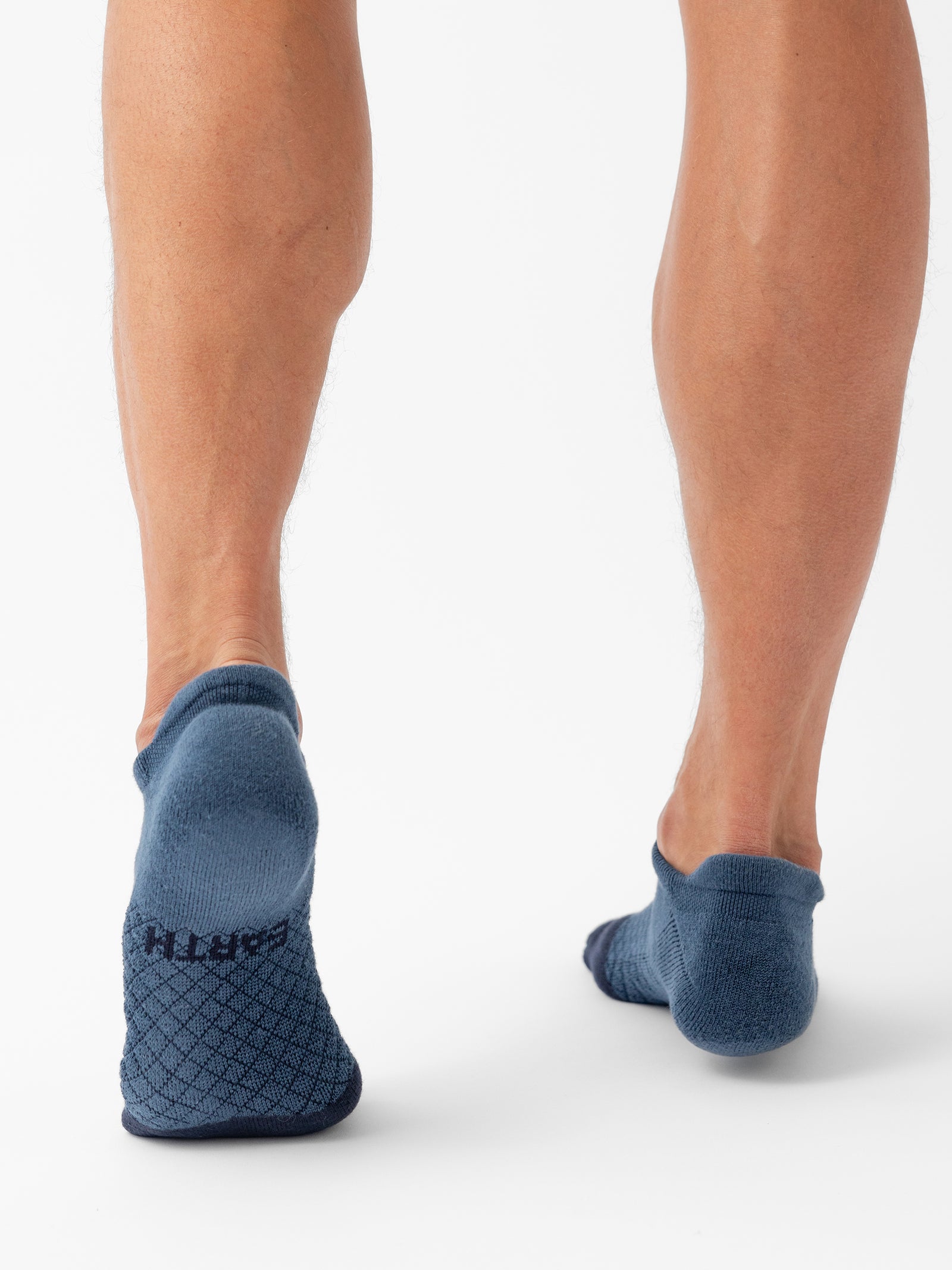 The image shows the lower legs of a person standing on tiptoes, wearing blue Cozy Earth Essential Ankle Socks with a textured pattern and the word "EARTH" printed on the back of one sock. The background is plain white. 