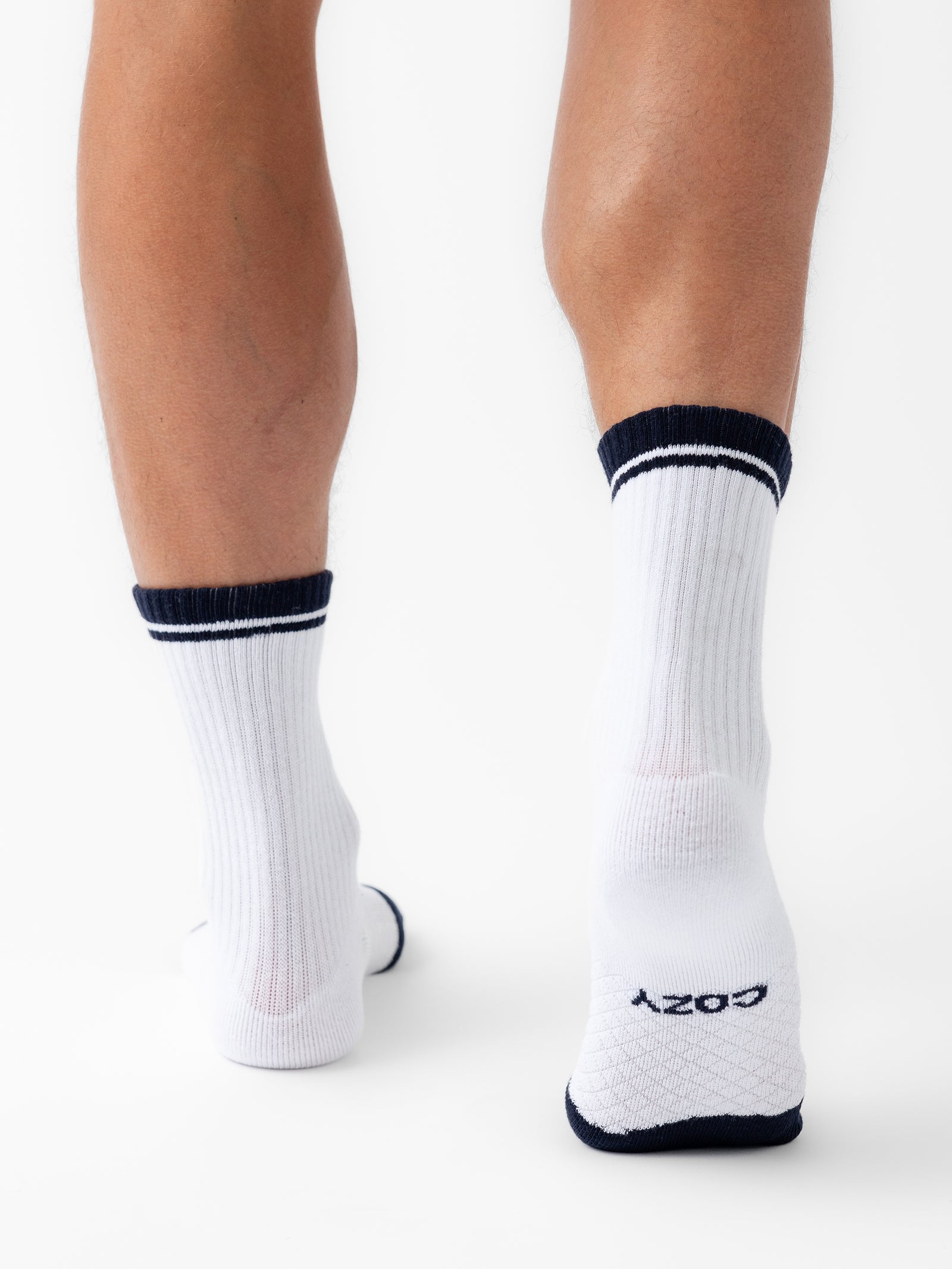 Close-up of a person's lower legs wearing white Essential Calf Socks from Cozy Earth, featuring black bands at the top and black soles. The back of the socks display "COZY" in black near the heel. The background is plain white. 