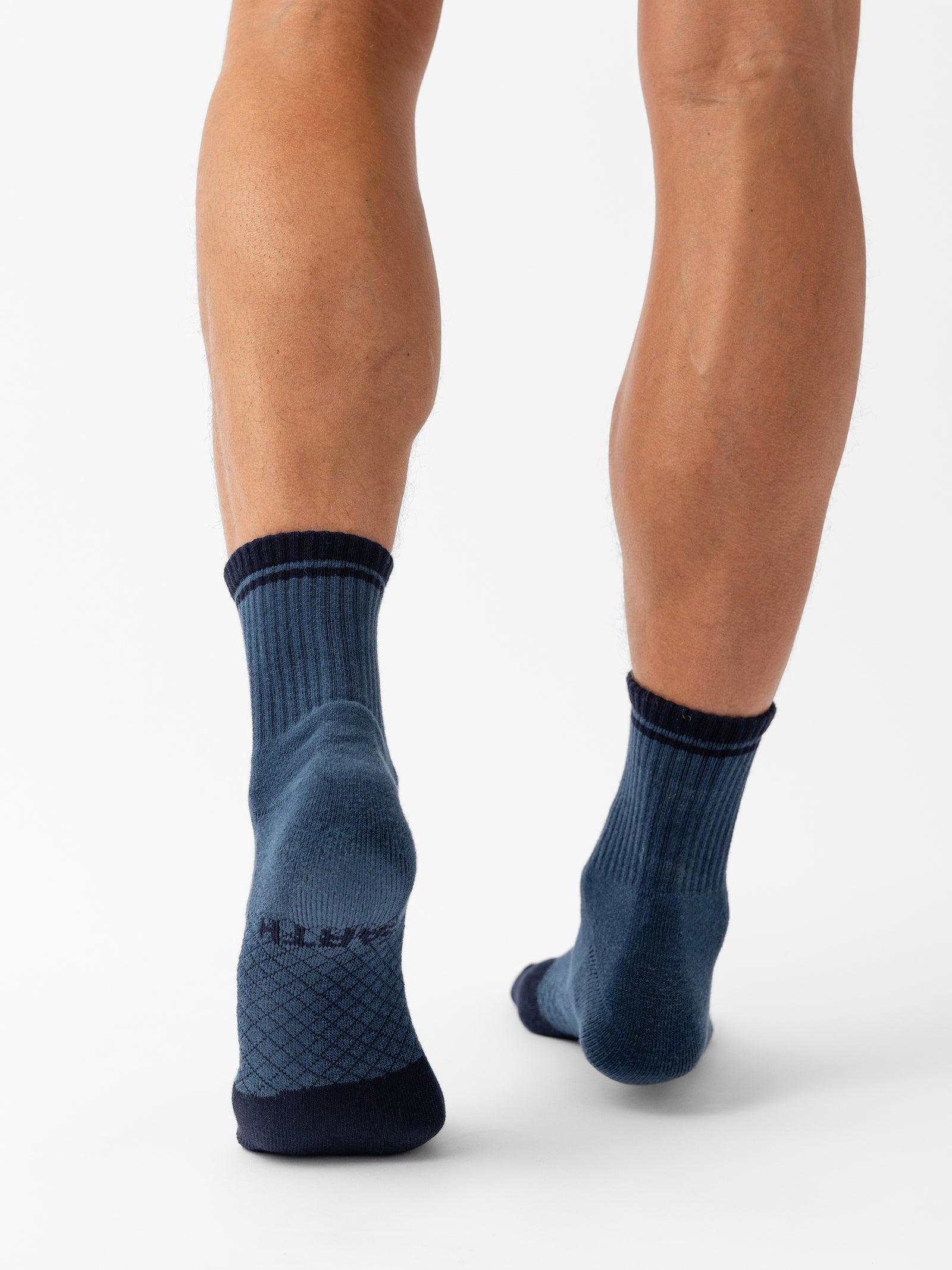 A person standing on a white background wearing a pair of Cozy Earth Essential Quarter Socks in blue. The socks feature a ribbed design at the top, dark blue toes and heels, and a textured pattern at the arches. Only the lower legs and feet of the person are visible. 