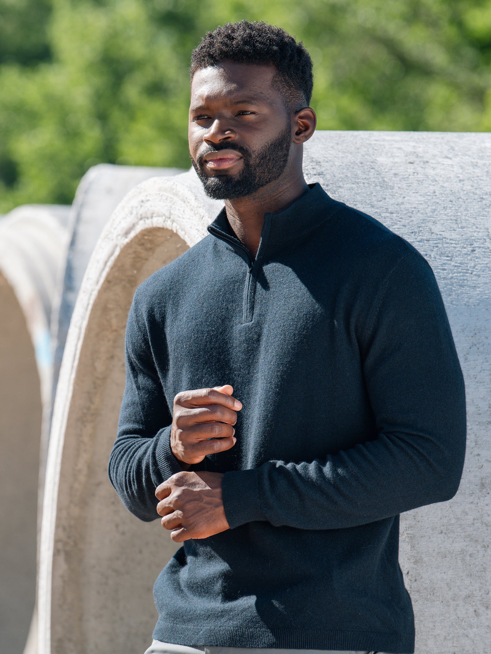 A man with a beard, wearing a dark long-sleeve Men’s Quarter Zip Sweater by Cozy Earth, stands near a large concrete structure. He holds his hands together and looks off into the distance. The background features greenery, suggesting an outdoor setting. 