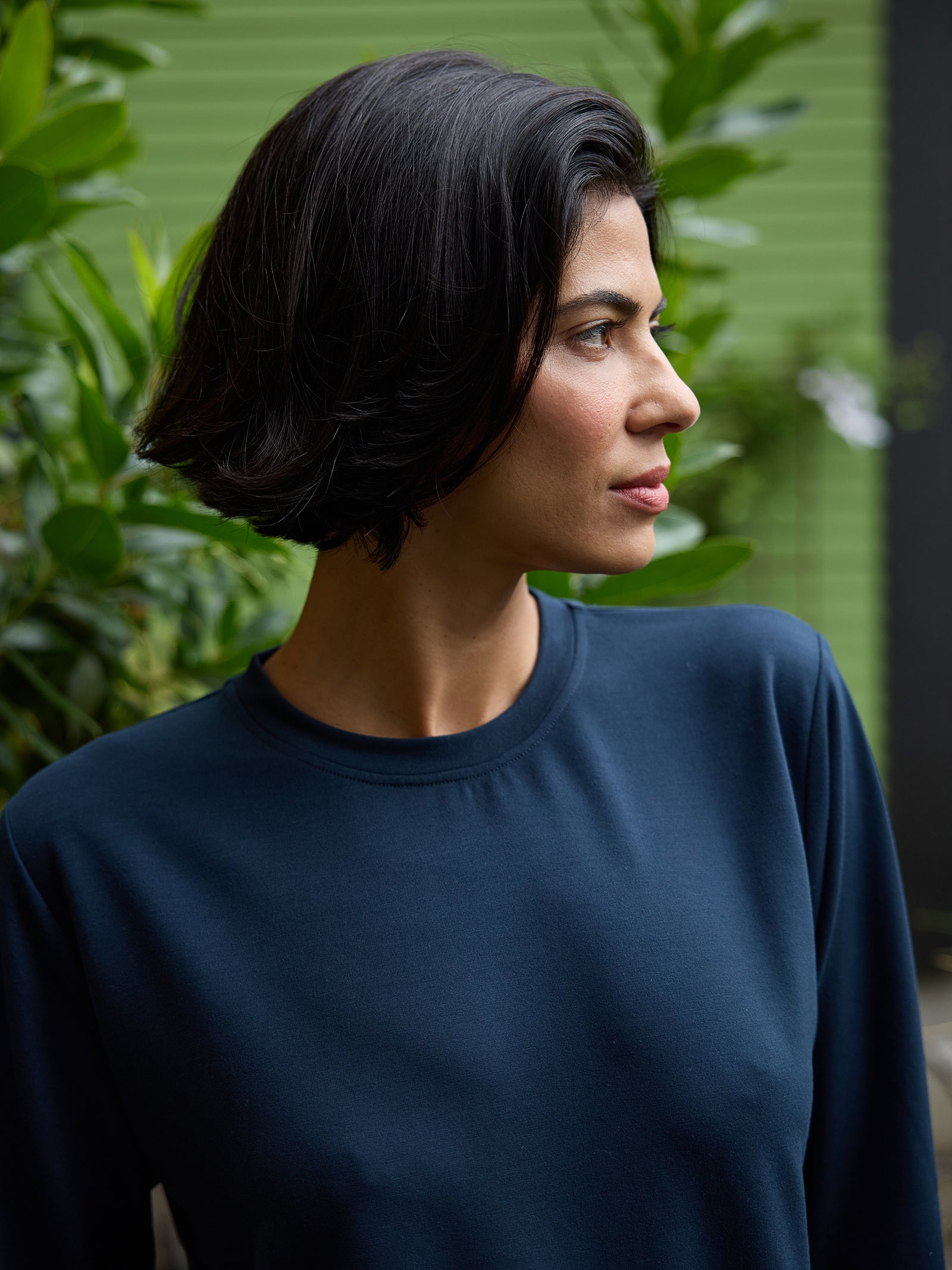 A woman with short, dark hair is looking to her right. She is wearing a Cozy Earth Women's StudioLite Crewneck in dark blue and is standing in front of a green, leafy background. The expression on her face appears contemplative. 