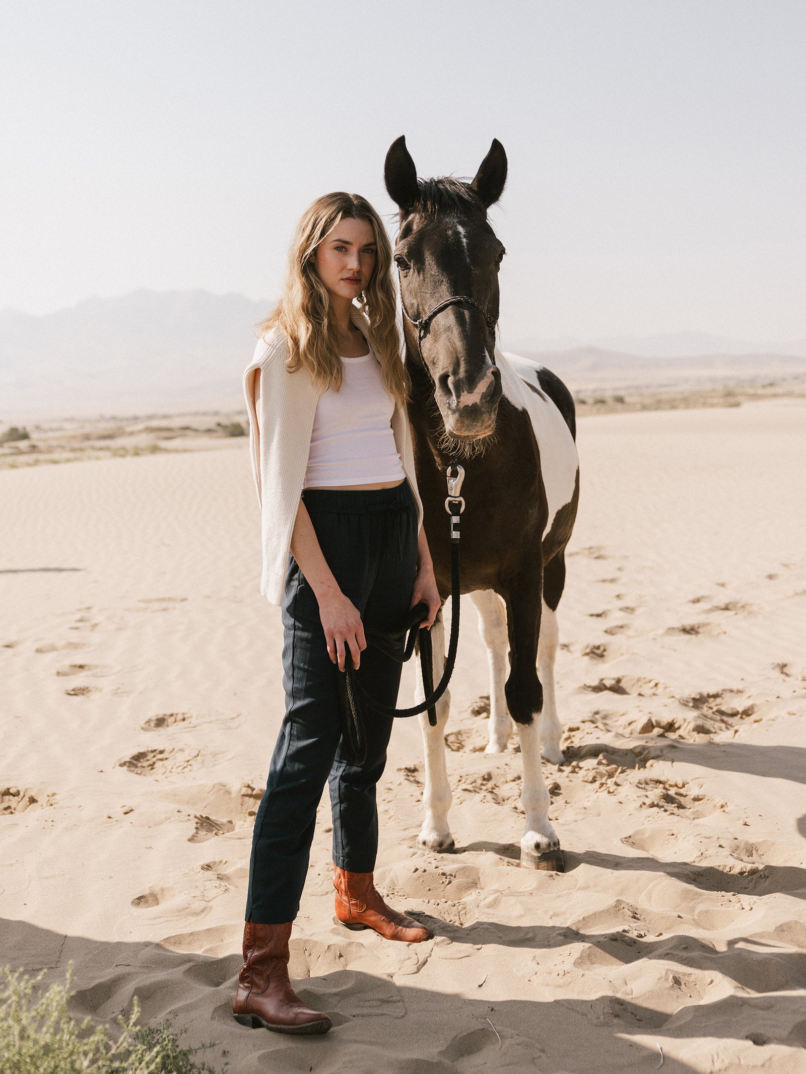 A person with long hair stands on a sandy landscape holding the reins of a black and white horse. The person is wearing a white top, Cozy Earth's Women's Sunset Cropped Pant, and brown boots, with a beige sweater draped over their shoulders. Desert mountains are visible in the background. 