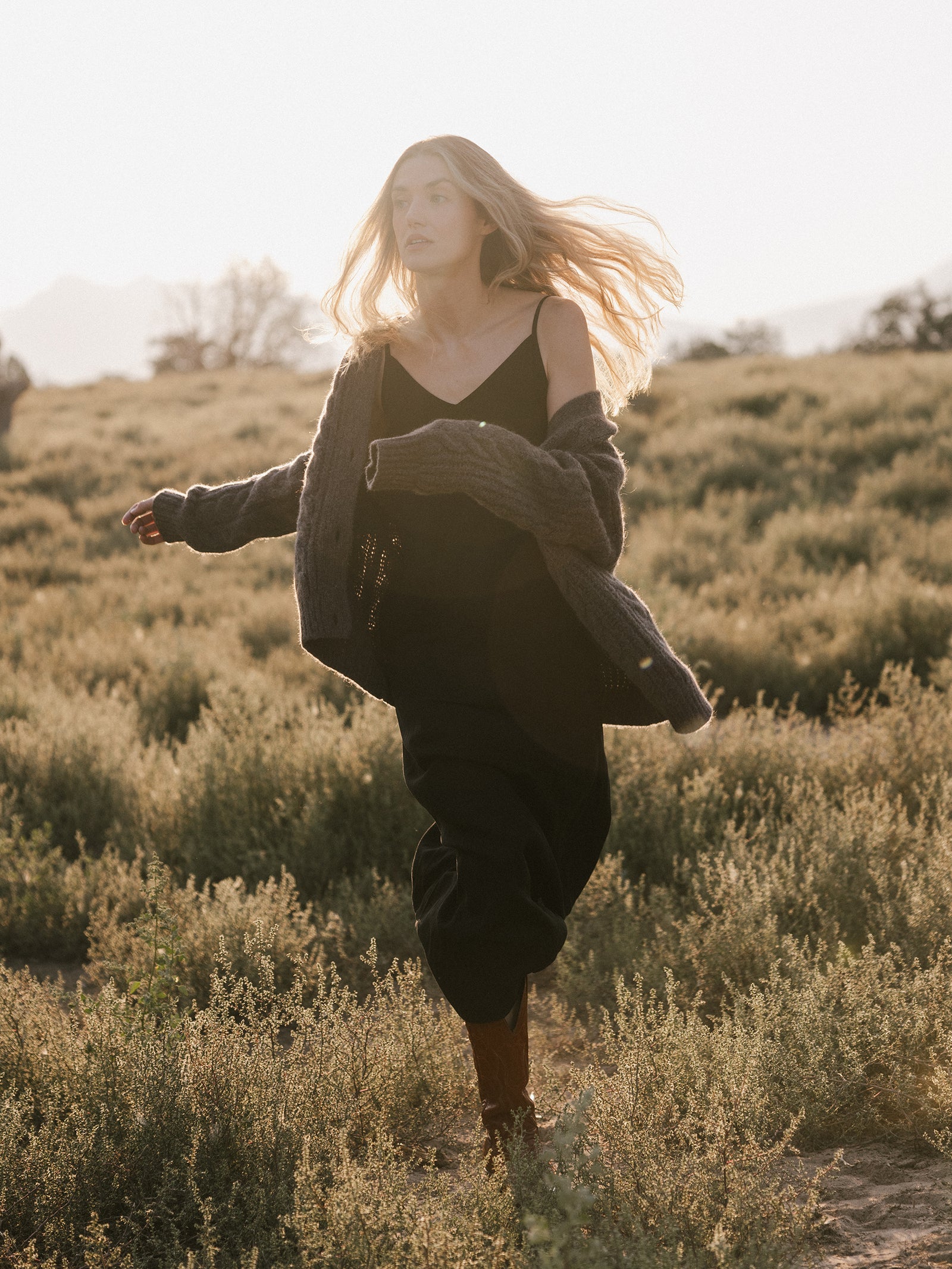 A woman with long hair walks through a grassy field during sunset. She is wearing the Women's Sunset Slip Dress from Cozy Earth, paired with a large, cozy sweater that is partially off her shoulders. Her hair flows in the breeze as she gazes forward, creating a serene and peaceful scene. 