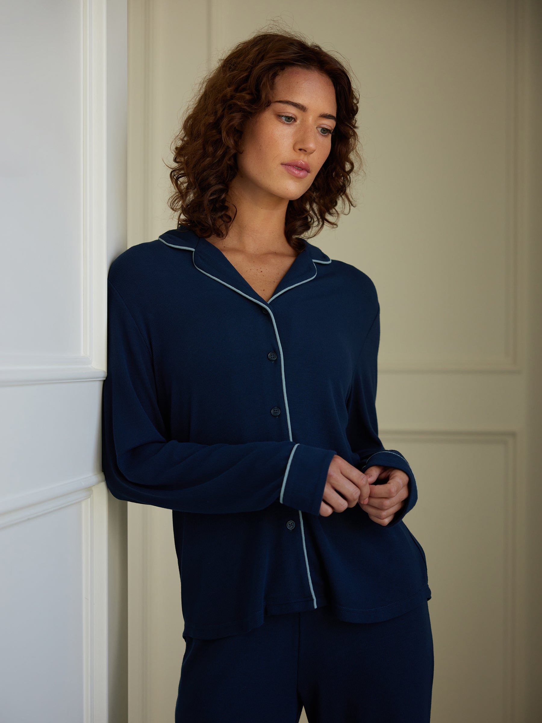 A person with curly hair stands pensively indoors, leaning against a white wall. They are wearing the Cozy Earth Women's Bamboo Rib Knit Classic Long Sleeve Pajama Top in dark blue, featuring light piping on the collar and cuffs. Their gaze is slightly downward and their hands are loosely clasped in front of them. 