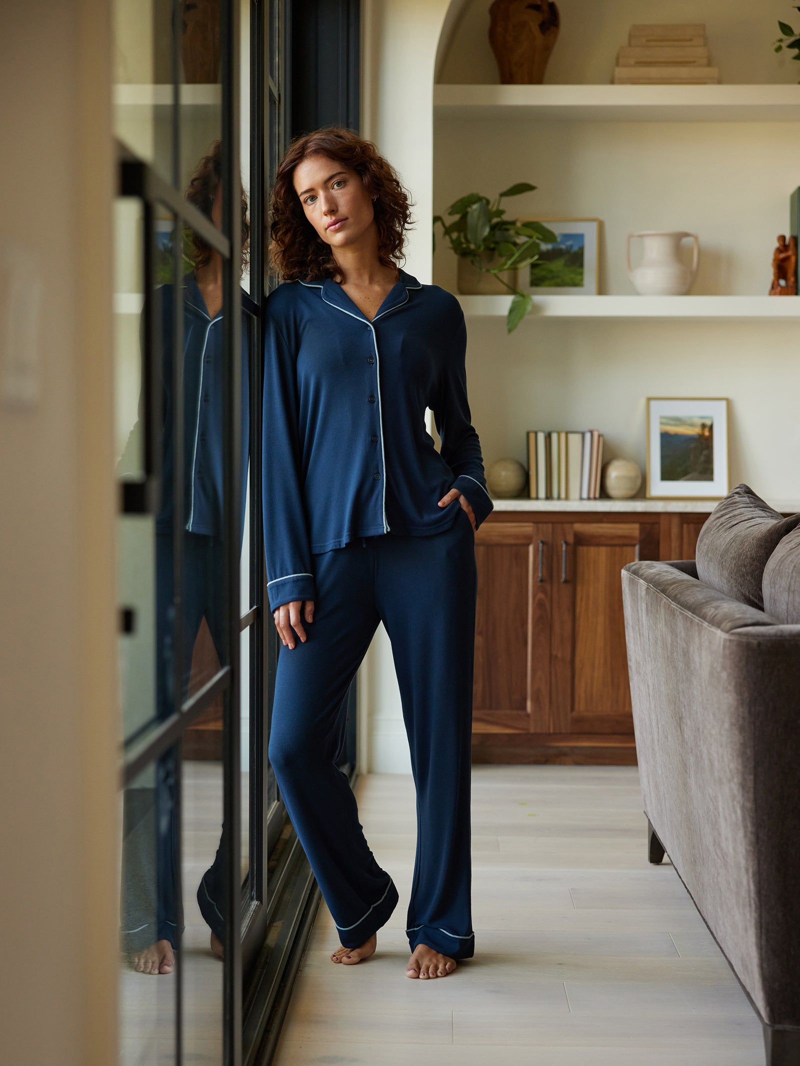 A woman with curly hair, dressed in Cozy Earth's Women's Bamboo Rib Knit Classic Pajama Pant in navy blue, stands barefoot by a large window in a cozy living room. She has her hands in her pockets and gazes thoughtfully at the camera. The room features a wooden cabinet, books, and potted plants. 