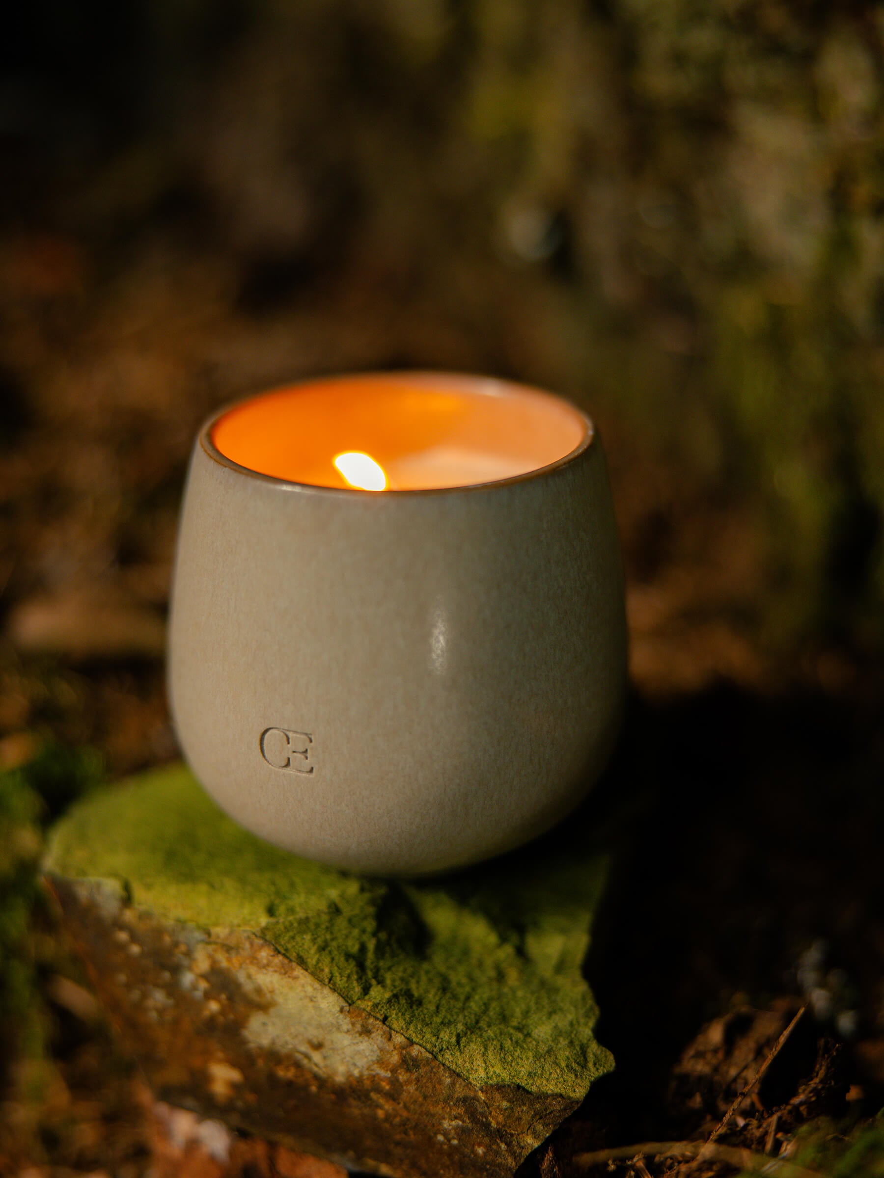 The Candle EMBER by Cozy Earth, in a sleek gray ceramic holder, sits on a mossy rock and casts a warm glow in a serene outdoor setting.