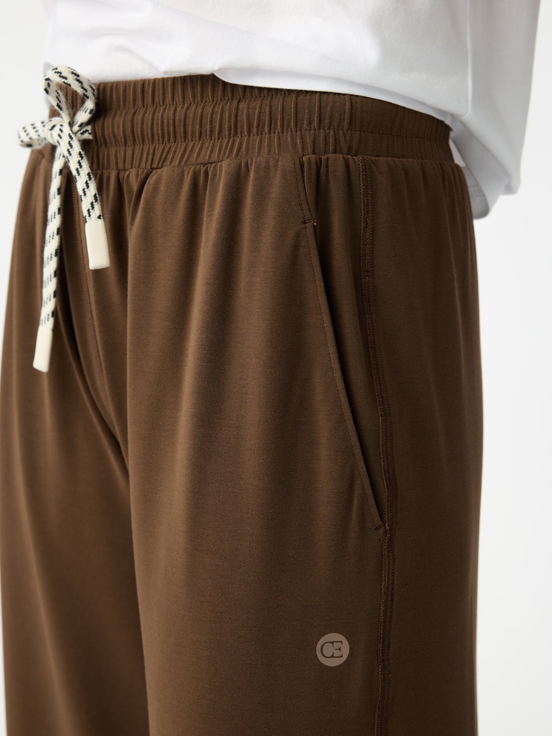 A person wearing Cozy Earth's Women's Studio Cropped Wide Leg Pant in brown, featuring a white-and-black drawstring and a small emblem near the pocket, paired with a white top. 