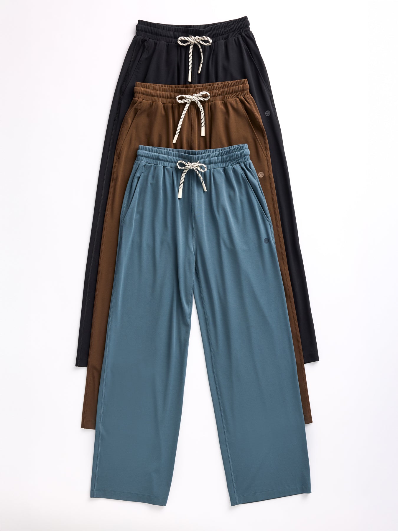 Four pairs of Cozy Earth's Women's Studio Wide Leg Pants in black, brown, and blue are stacked neatly on a white background. Each features an elastic waistband with a white drawstring. 