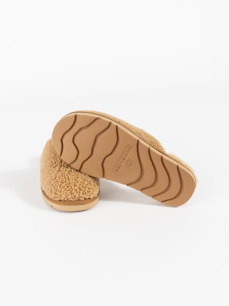 A pair of Cozy Earth's Puffy Sheep Slippers, featuring a tan fleece texture and embossed rubber soles with a wavy grip pattern, displayed in a studio setting. The slippers are arranged to showcase both the top and bottom views.
