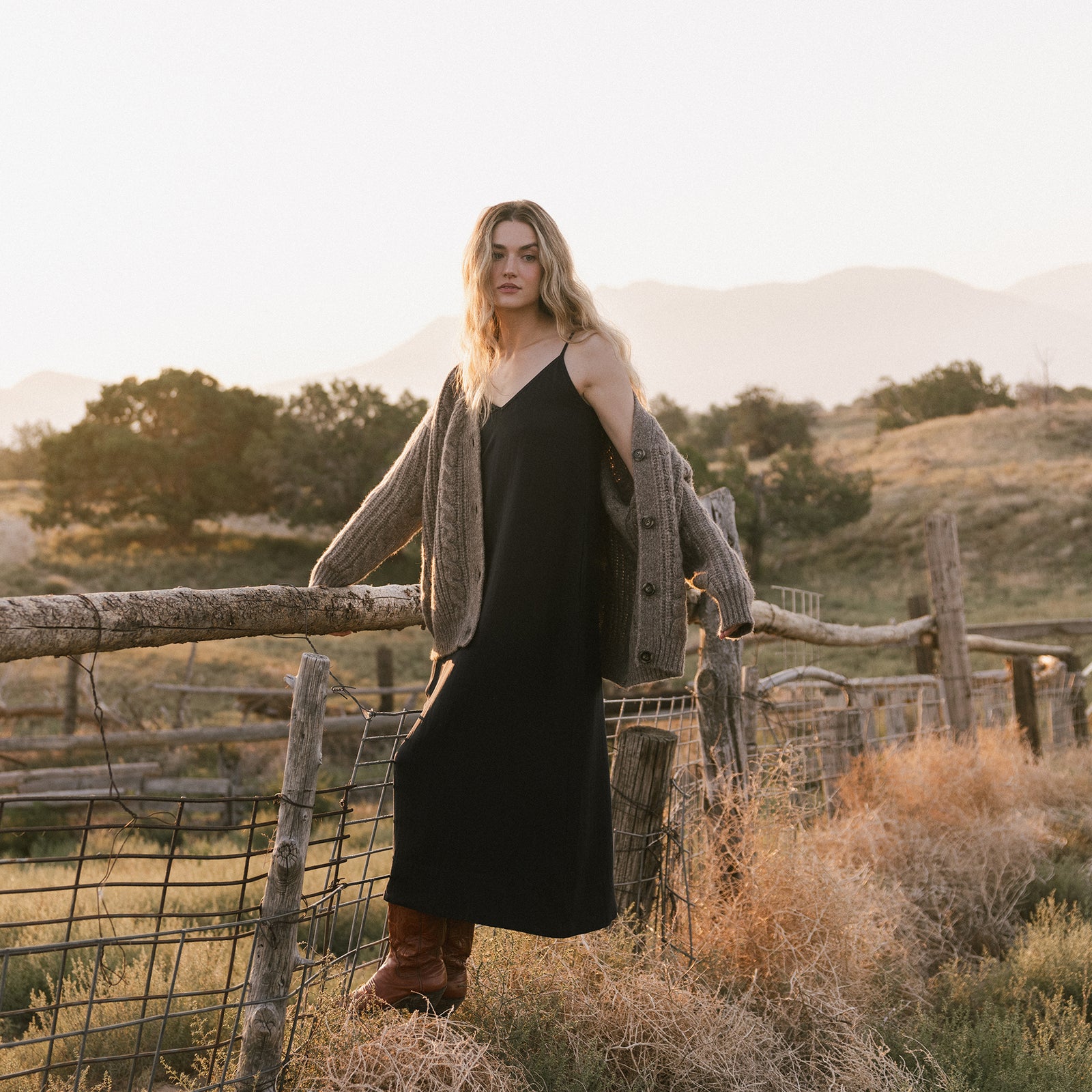 A person with long hair stands on a rustic fence in a rural setting, wearing a long black dress, an Oversized Cable Knit Cardigan by Cozy Earth, and brown boots. The background features rolling hills and sparse trees under a softly lit sky. 