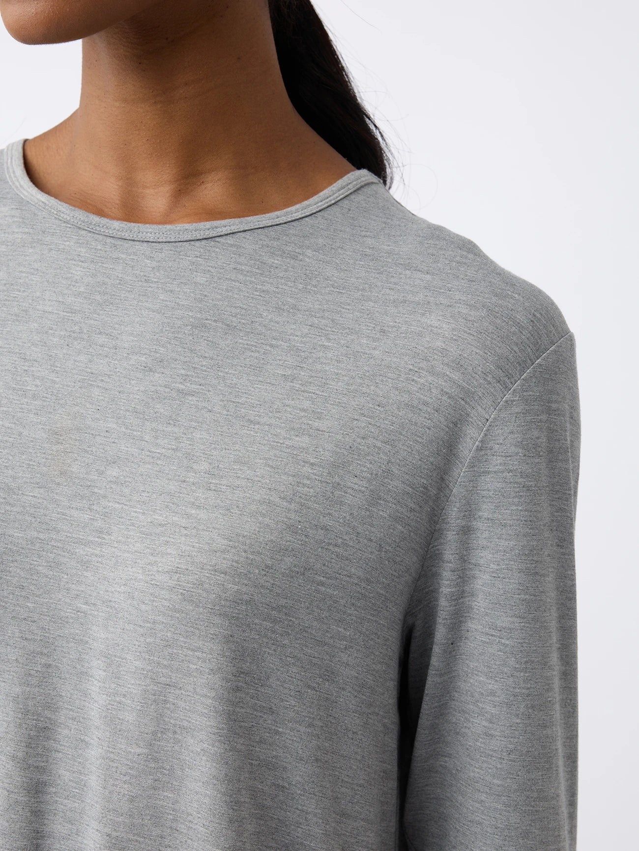 A woman wearing the Cozy Earth Women's Stretch Knit Bamboo Long Sleeve Lounge Tee in light gray stands against a plain background, showing her shoulder and part of her face with hair tied back. 