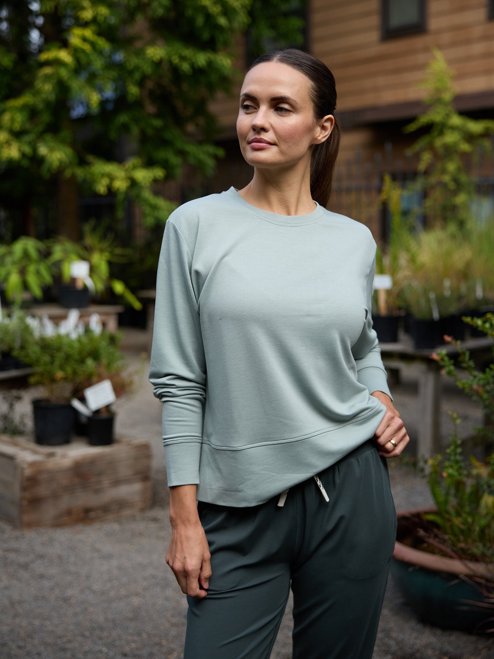 A woman stands outdoors in a garden center wearing a Cozy Earth Women's StudioLite Crewneck and dark gray sweatpants. She is looking to the side with her hand resting on her hip, surrounded by plants and small trees in black pots. In the background, there is a wooden building. 