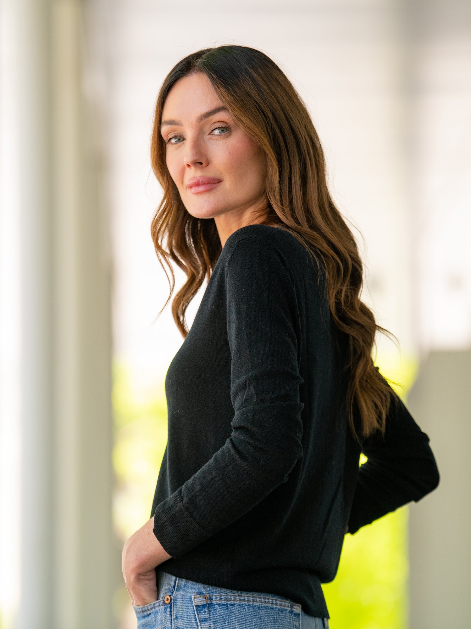 A woman with long, wavy brown hair stands sideways, looking at the camera with a slight smile. She is wearing a Cozy Earth Women's AirKnit V-Neck Sweater in black and blue jeans. The background is bright, with blurred green and white elements. 