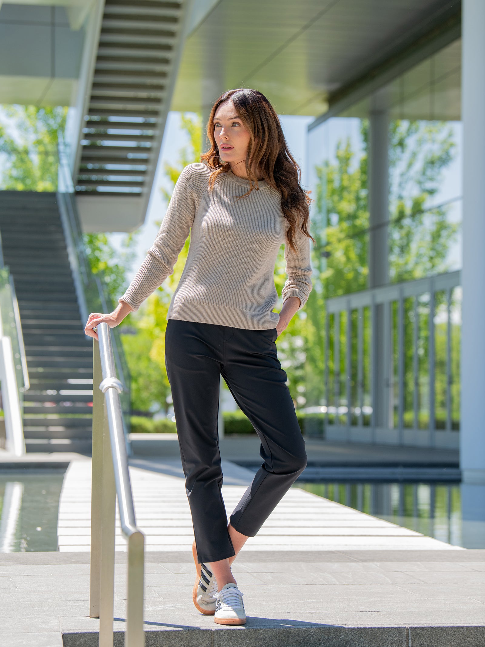 A woman with long hair stands outdoors near a staircase, leaning on a handrail. She is wearing a beige sweater, Cozy Earth's Women's Always Cropped Pant in black, and gray sneakers. The background features modern architecture with glass walls and greenery. 