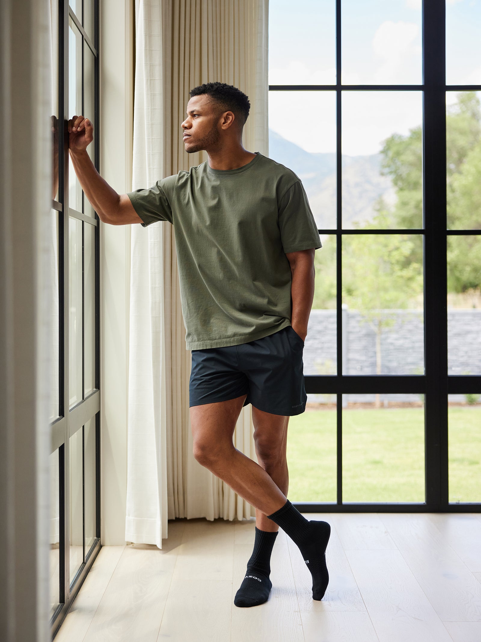 A man wearing a green t-shirt, Cozy Earth's Men's Performance Sleep Short in black, and black socks stands by a large window, resting one arm on the window frame and looking thoughtfully outside. The room is well-lit with light wooden floors and offers a view of greenery and mountains beyond. 