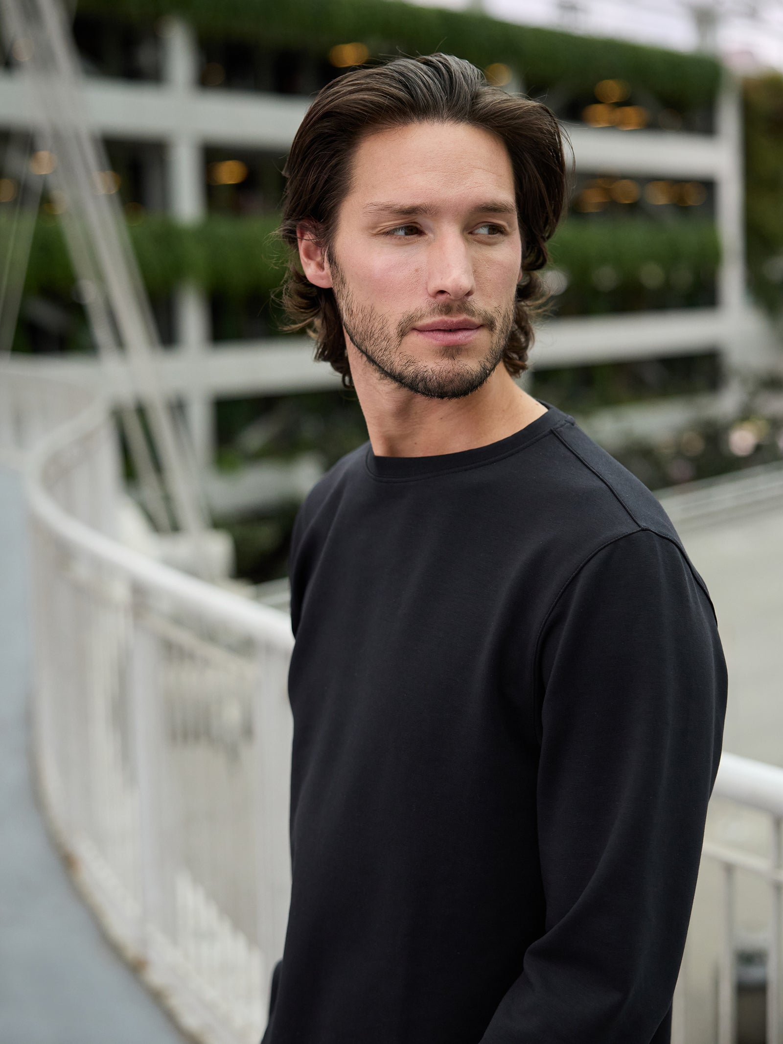 A man with shoulder-length brown hair and a beard is wearing a black Cozy Earth Men's StretchTech Crewneck long-sleeve shirt. He stands near an outdoor railing and looks to his left. The background features a multi-level structure with greenery and industrial elements. 