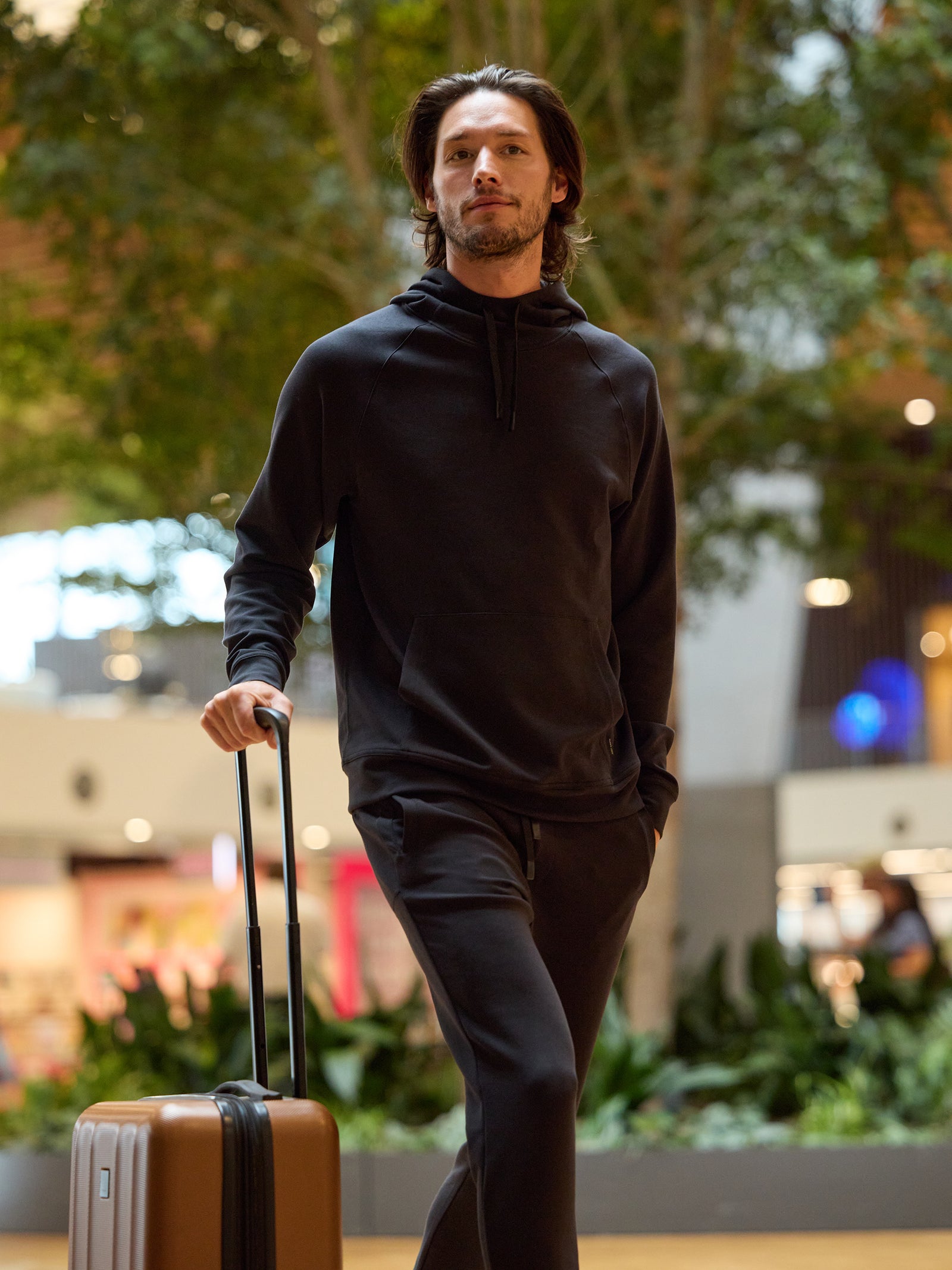 A man with shoulder-length hair walks through an indoor setting, such as an airport or mall, pulling a suitcase with one hand. He is wearing Cozy Earth's Men's StretchTech Hoodie and matching black pants. The background features greenery and blurred lights. 