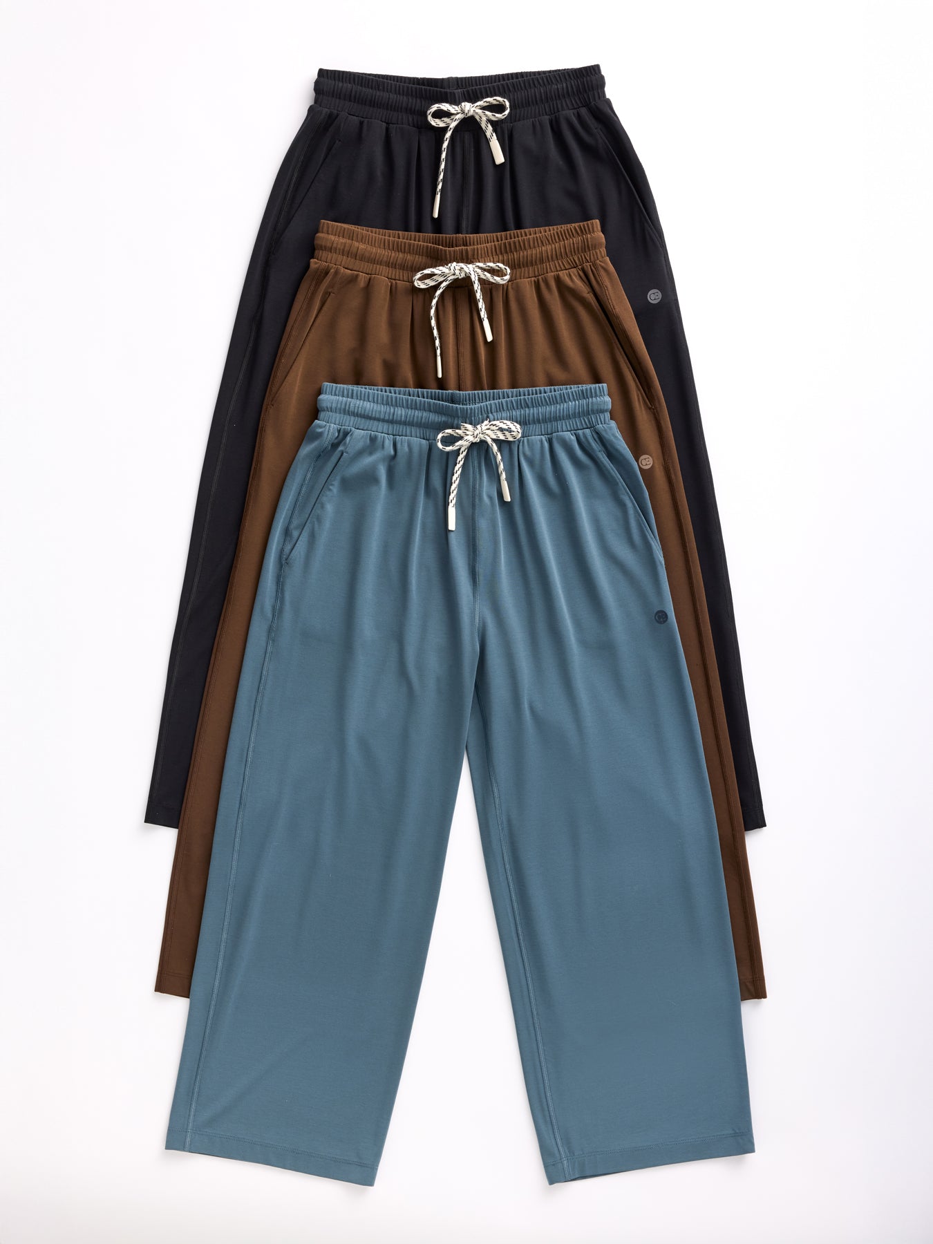 Four pairs of Cozy Earth's Women's Studio Cropped Wide Leg Pants with a drawstring waist and side pockets are stacked. From top to bottom, the colors are black, brown, and light blue. 