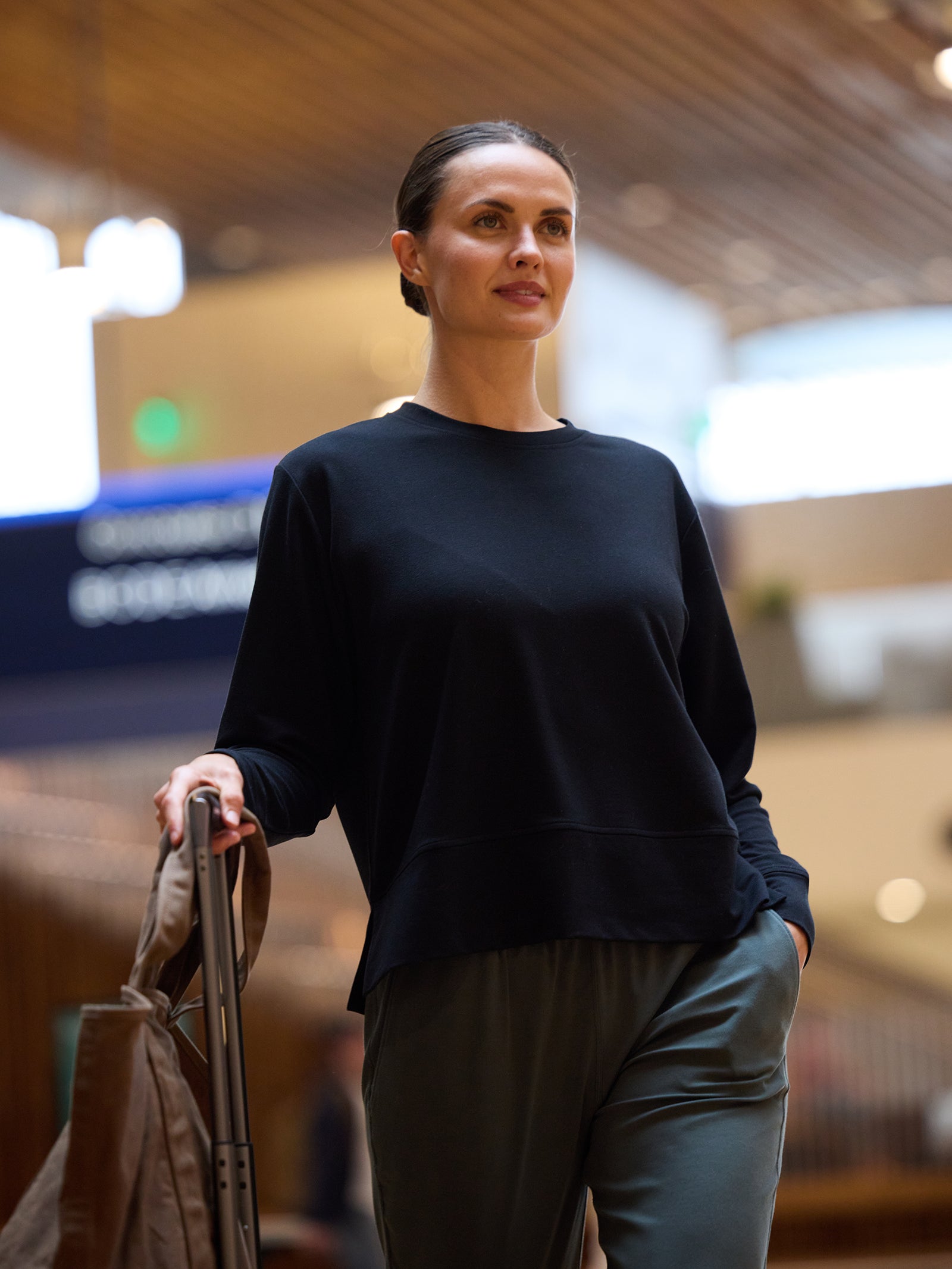 A woman with a bun hairstyle, wearing the Cozy Earth Women's StudioLite Crewneck in black along with green pants, strolls confidently through an indoor space while holding a rolling suitcase. The blurred background features modern architecture with wooden elements and a lit sign. 