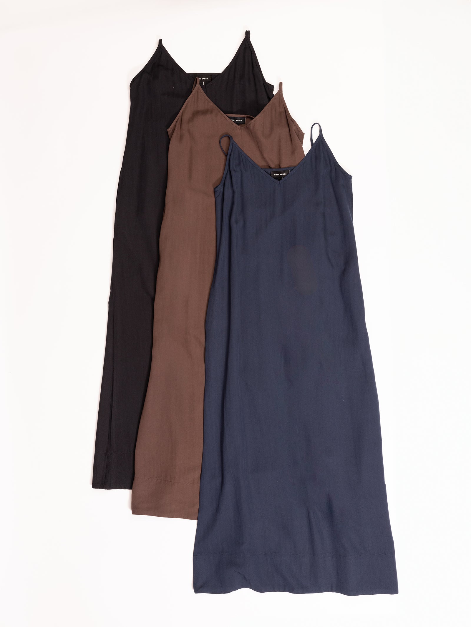 Three Women's Sunset Slip Dresses by Cozy Earth are laid out in a staggered arrangement. The dresses are of the same design but come in three different colors: black, brown, and navy blue. Each dress features a V-neckline and thin shoulder straps. The background is plain white. 