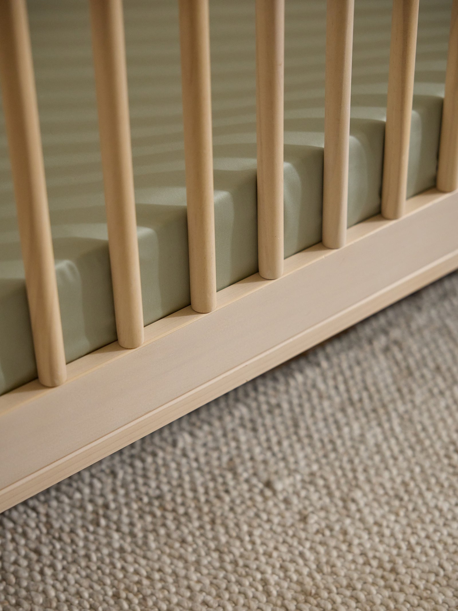 Close-up view of a light wooden crib rail and slats with the Cozy Earth Crib Sheet by Cozy Earth. The crib rests on a textured beige carpet, creating a calm and cozy atmosphere. 