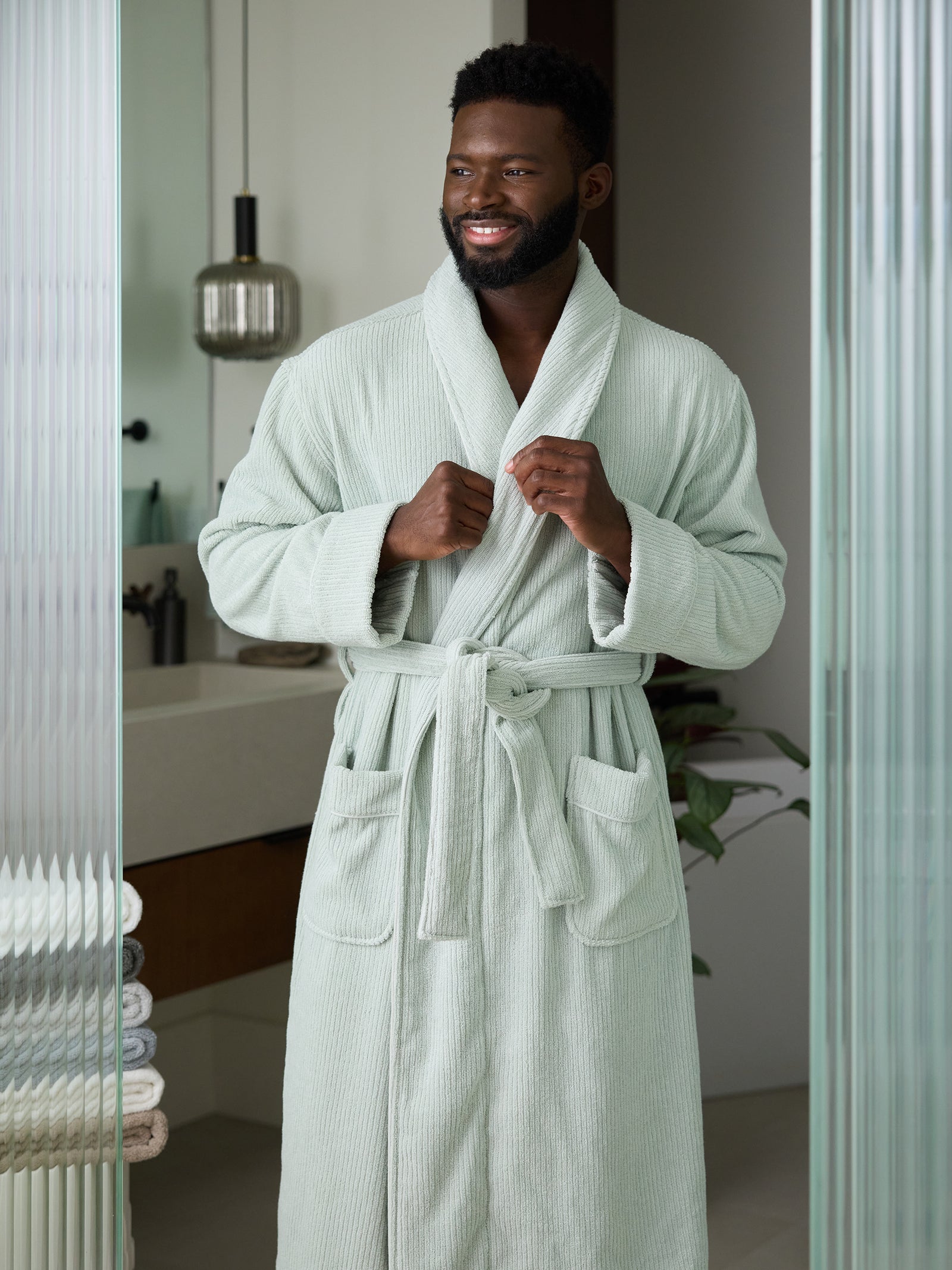 A man stands smiling in a bathroom, wearing a Cozy Earth Ribbed Terry Bath Robe, characterized by its light green color, tie belt, and two front pockets. In the background, there is a sink, a stack of towels, and a modern hanging light fixture. The setting appears clean and well-lit. 