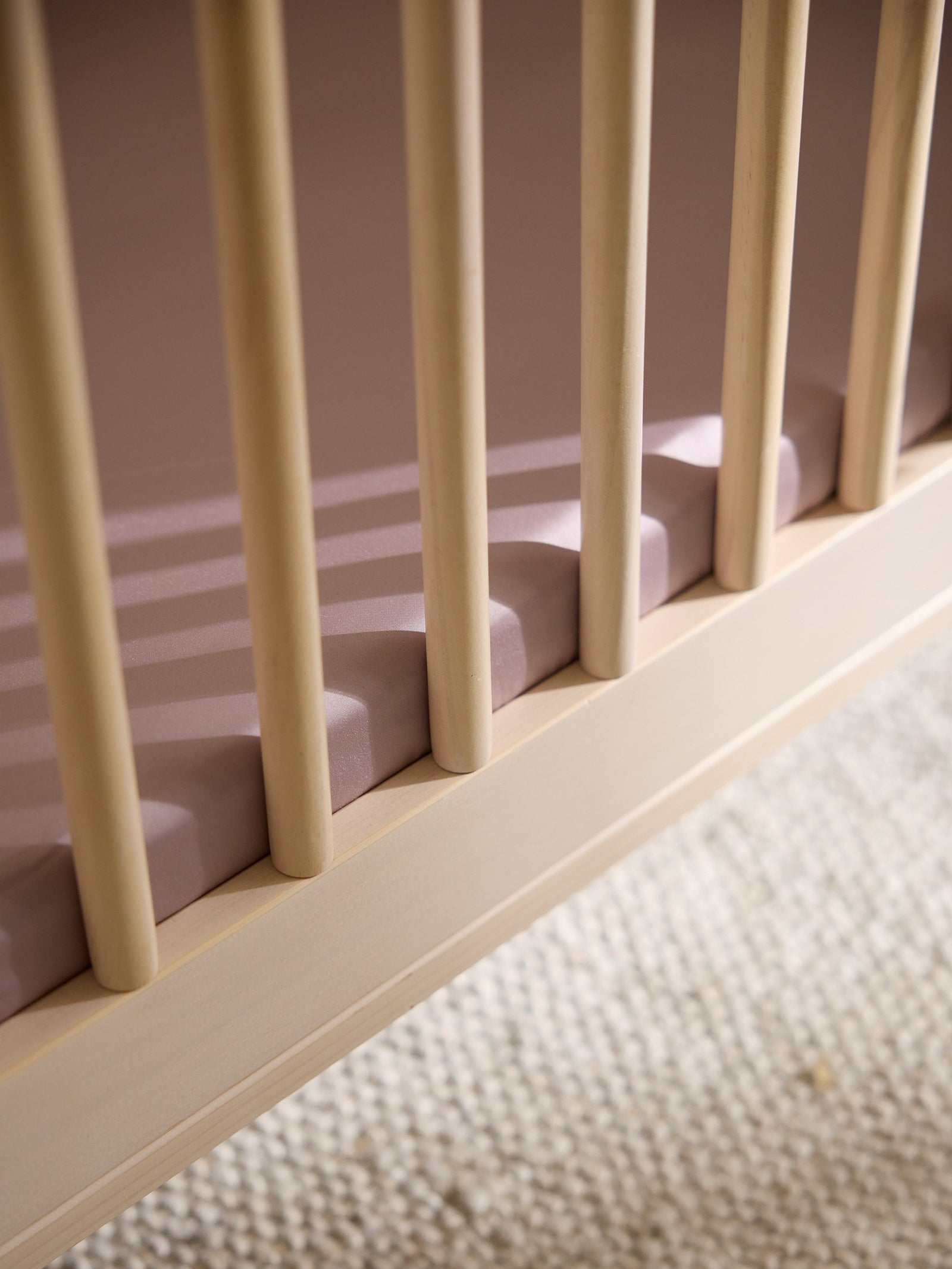 Close-up of a wooden crib rail with vertical slats casting shadows on a pink Cozy Earth Crib Sheet by Cozy Earth, placed on a textured beige carpet. 