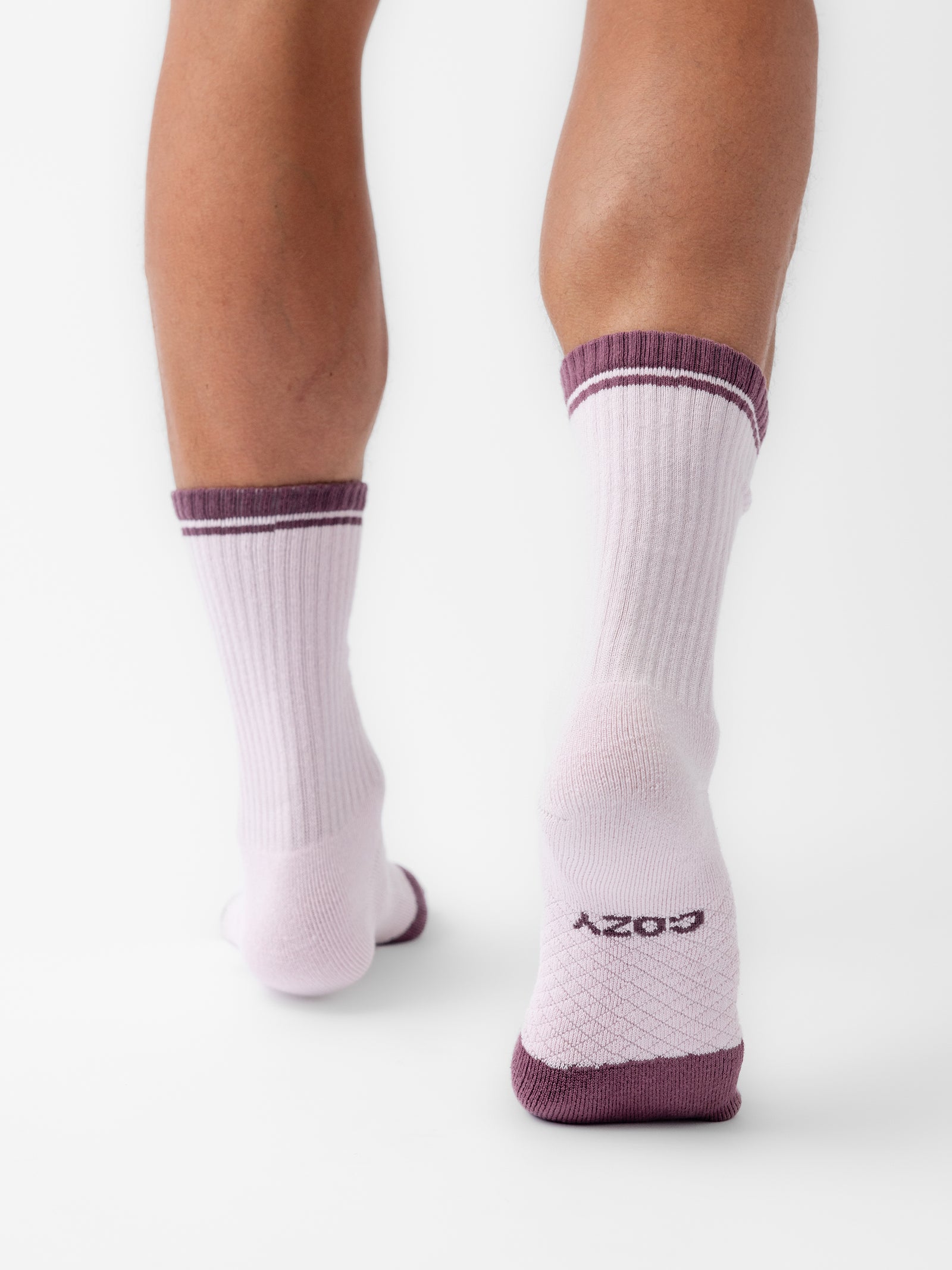 The image shows a close-up of a person's lower legs and feet from behind, wearing a pair of white Essential Calf Socks with ribbed design and dark purple trim at the top, featuring the word "COZY" written on the soles in purple. The person is standing on a white background. These socks are part of the Cozy Earth Essential Calf Sock 2-Pack. 