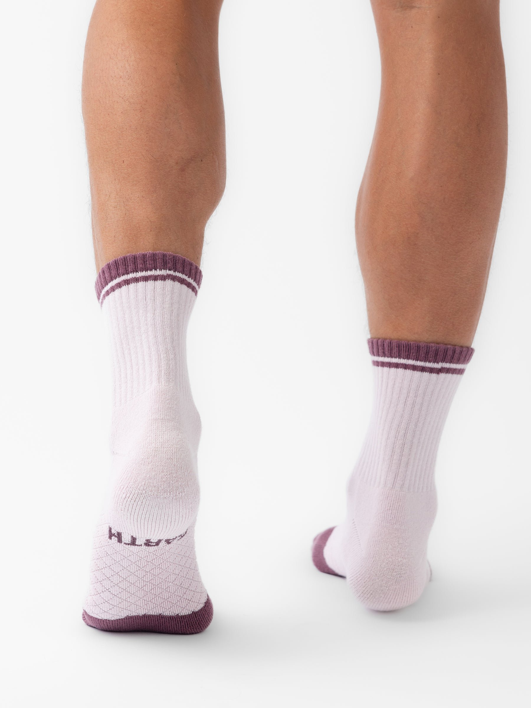 The image displays the back view of a person's legs wearing Cozy Earth's Essential Quarter Socks in white with purple trim. The socks feature ribbed patterns and a honeycomb texture on the sole, a purple stripe near the top, and "Cozy Earth" visible at the heel. 