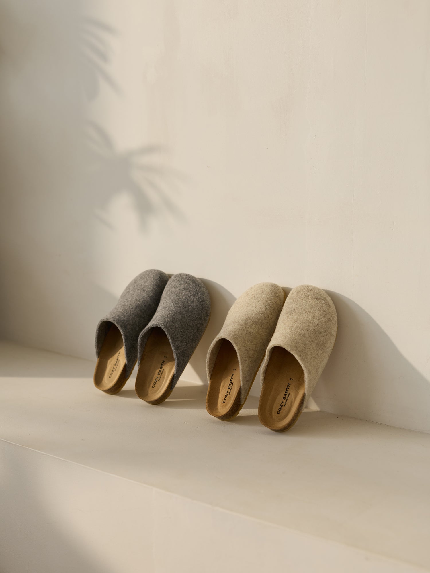 Two pairs of Cozy Earth's Lakehouse Clogs sit on a light surface. One is gray, the other beige, with soft sunlight casting gentle shadows of leaves on the wall behind them, creating a calm and cozy ambiance.