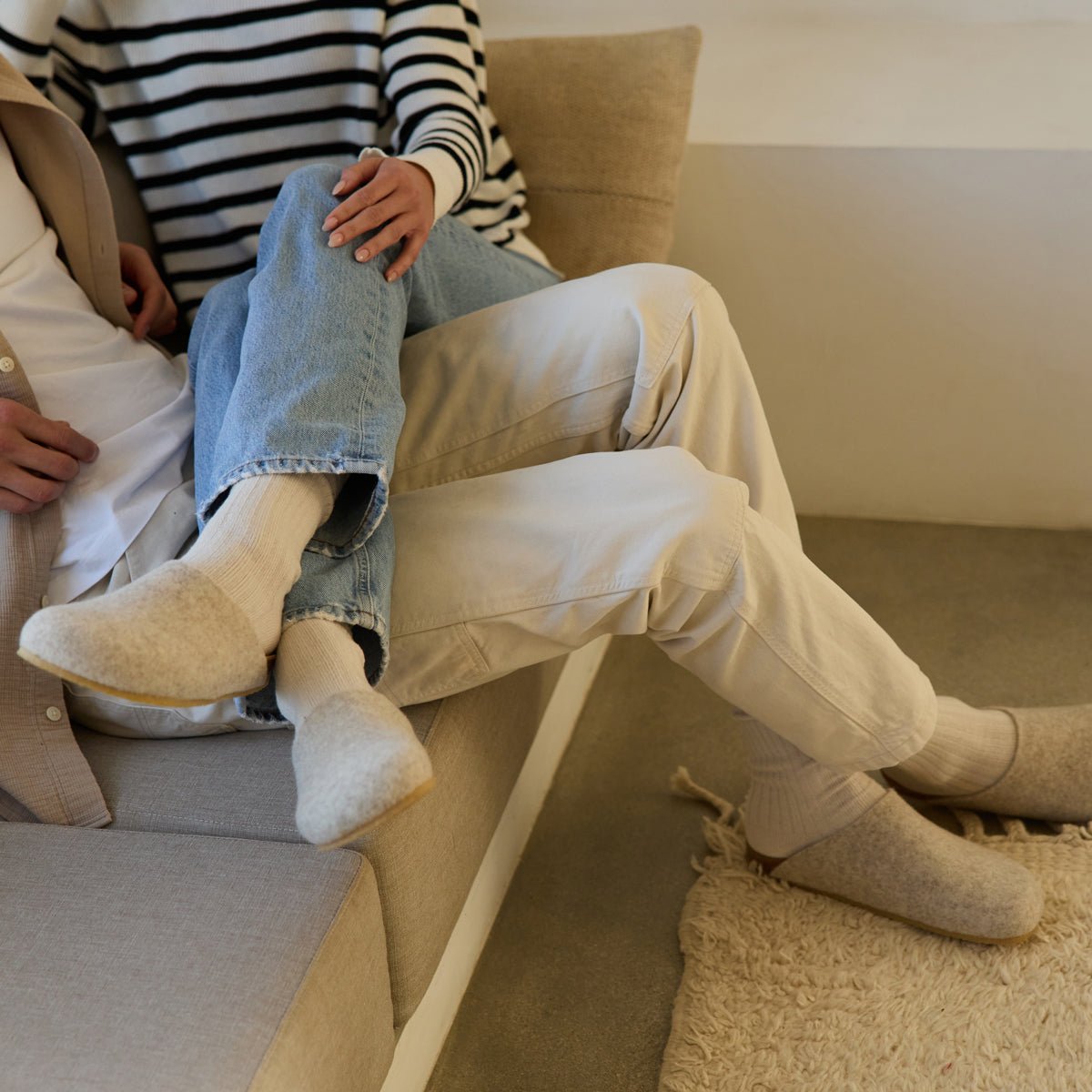 Two people relax on a beige sofa in casual, light-colored clothing. One in striped tops and jeans sits across the other's lap. Both wear Cozy Earth's Lakehouse Clog wool slippers, creating a cozy atmosphere. 